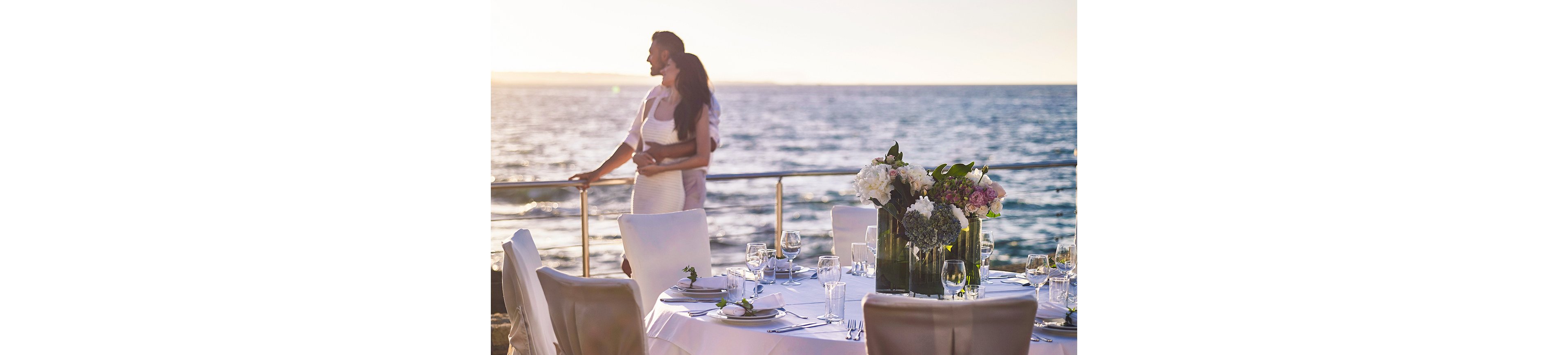 A couple stands embracing by a railing, gazing out over the ocean at sunset. In the foreground, an elegantly set round table is arranged for a formal meal with white linens, plates, glassware, and cutlery. Tall vases filled with white and pink flowers serve as centerpieces, adding a refined touch to the outdoor setting. The soft evening light and calm water create a romantic, celebratory atmosphere.