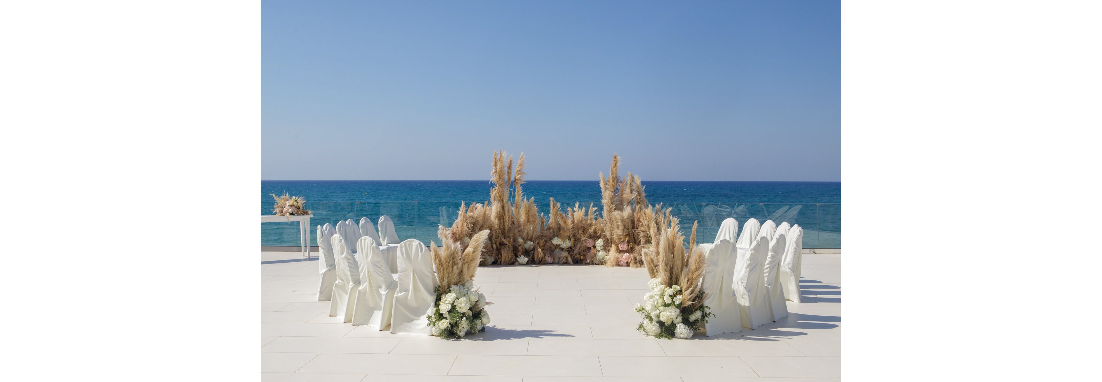 An outdoor wedding ceremony is arranged on a white terrace overlooking a calm blue sea. White-covered chairs form a semi-circle around an aisle, creating an intimate seating area. The focal point is a lush display of pampas grass and neutral-toned florals at the front, complemented by white flower arrangements near the chairs. The clear sky and ocean backdrop give the scene a serene, romantic atmosphere.