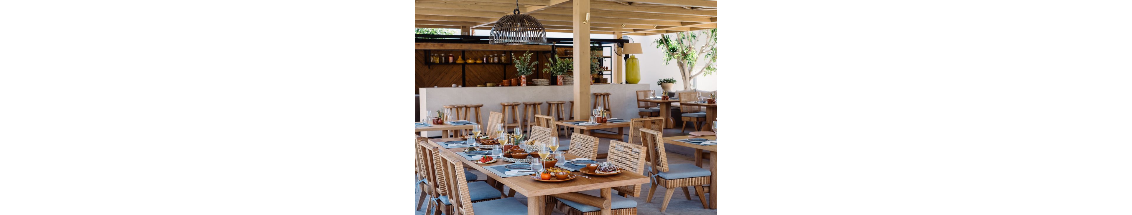 An open-air restaurant features neatly set wooden tables with woven chairs and blue seat cushions. Plates of food, wine glasses, and water glasses are arranged on the tables, ready for guests. In the background, a staff member works behind a bar counter decorated with plants, jars, and bottles. Natural light, neutral tones, and greenery create a relaxed, Mediterranean-inspired dining atmosphere.