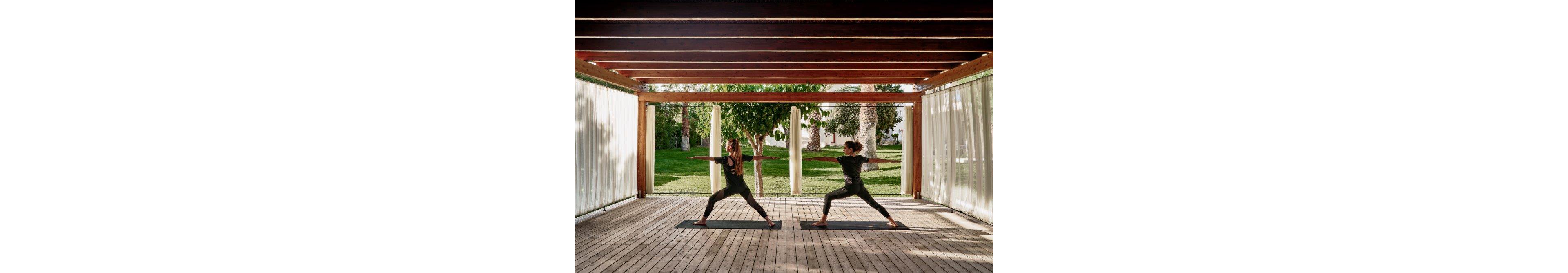 Two women perform yoga poses on mats in a wooden open-air pavilion. They stand facing each other in a wide stance, extending their arms in a balanced posture. Sheer curtains frame the structure, allowing natural light to filter in. A lush green lawn, trees, and a building in the background create a calm, outdoor retreat atmosphere.