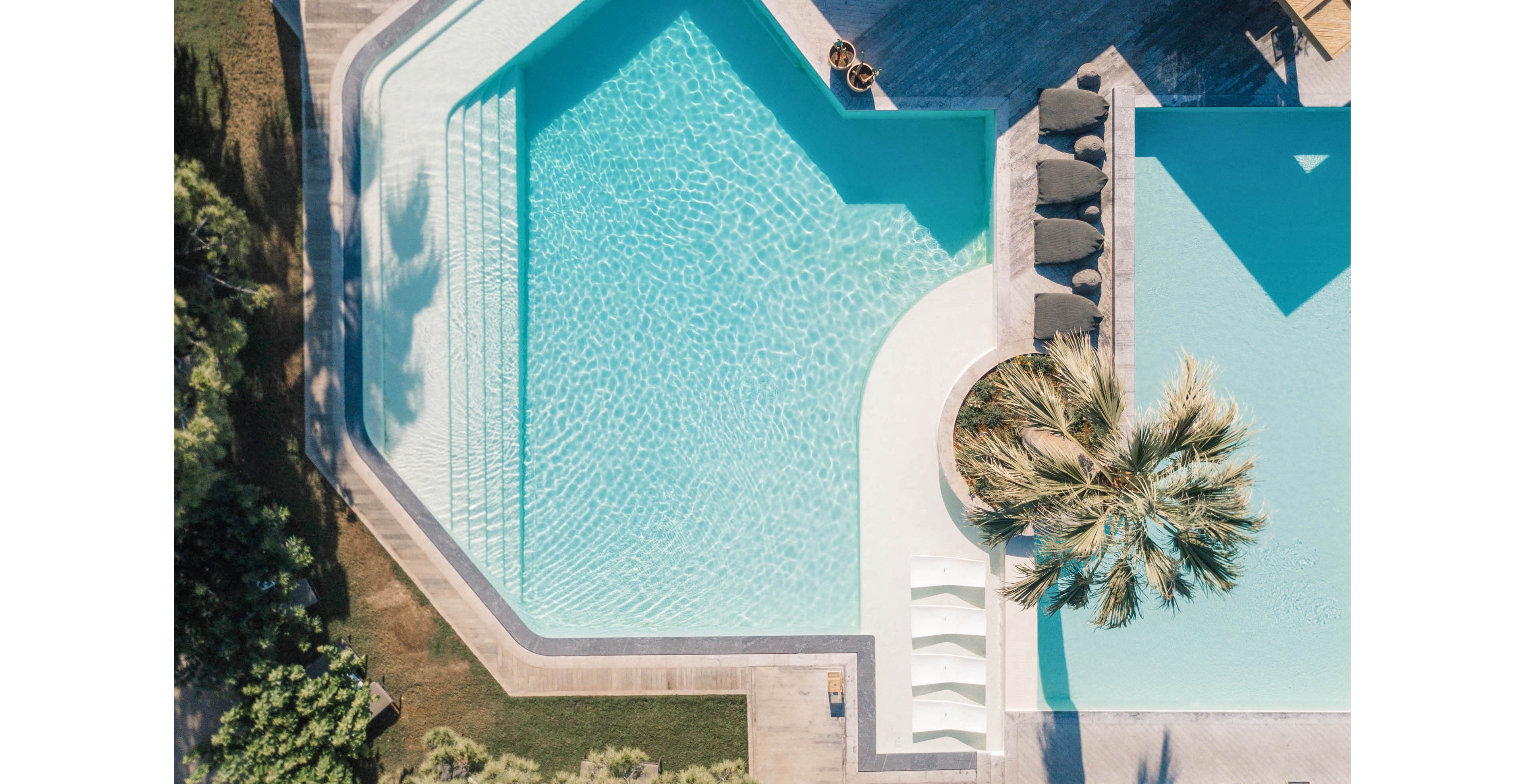 A single swimmer floats in a large geometric swimming pool captured from above. The pool is surrounded by minimalist concrete decking, sun loungers, and shaded seating areas. A tall palm tree and surrounding greenery add a tropical touch to the modern resort setting. Clear turquoise water, sharp lines, and soft shadows create a calm, luxurious vacation atmosphere.