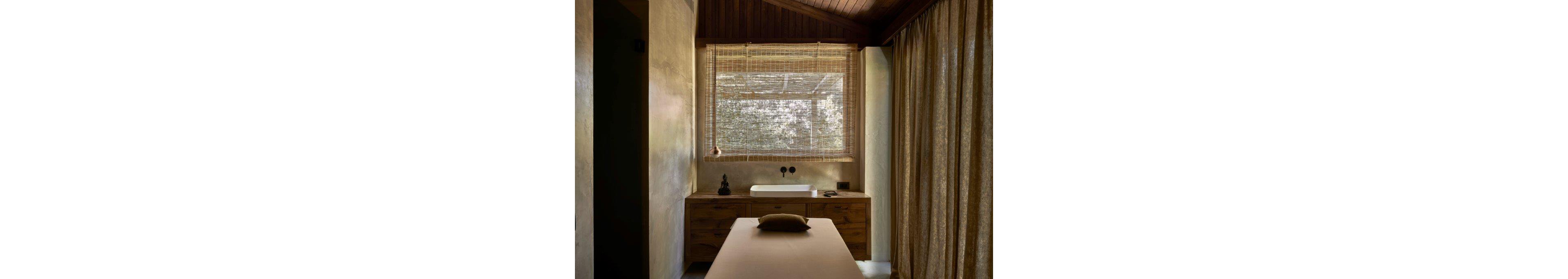 A tranquil spa treatment room features a massage table centered beneath a large window. Soft natural light filters through woven blinds, casting gentle shadows on the neutral-toned walls. A wooden vanity with a sink, small decor objects, and minimalist fixtures enhances the warm, organic feel. Floor-to-ceiling curtains add privacy and contribute to the cozy, relaxing atmosphere.