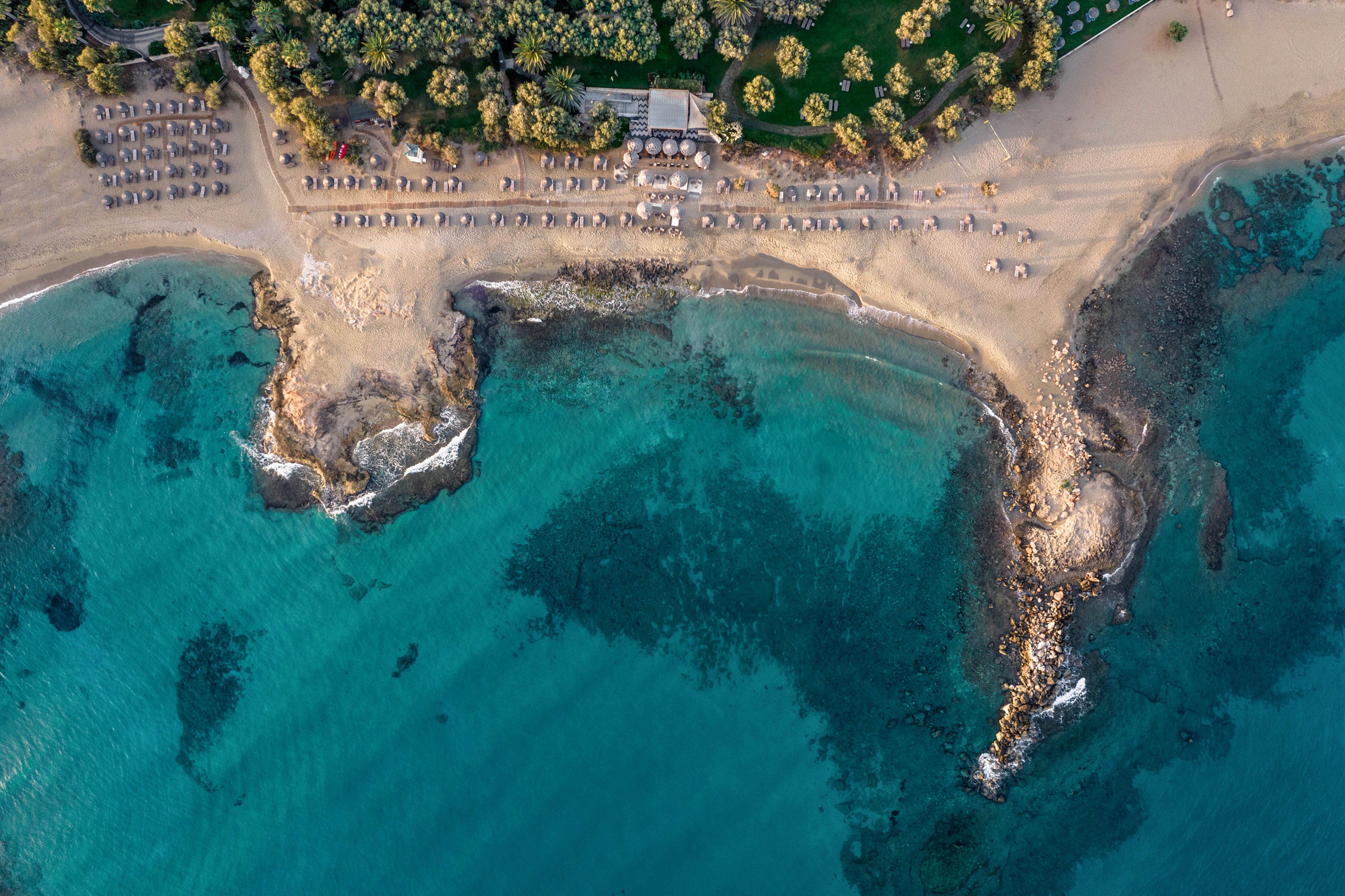 Aerial view of a tranquil sandy beach with clear turquoise water and rocky outcrops. Neatly arranged sun loungers and umbrellas line the shore near a small beachfront structure. The coastline curves gently, with shallow reefs visible beneath the calm sea. Lush green trees and a manicured lawn border the beach, creating a contrast between land and water.