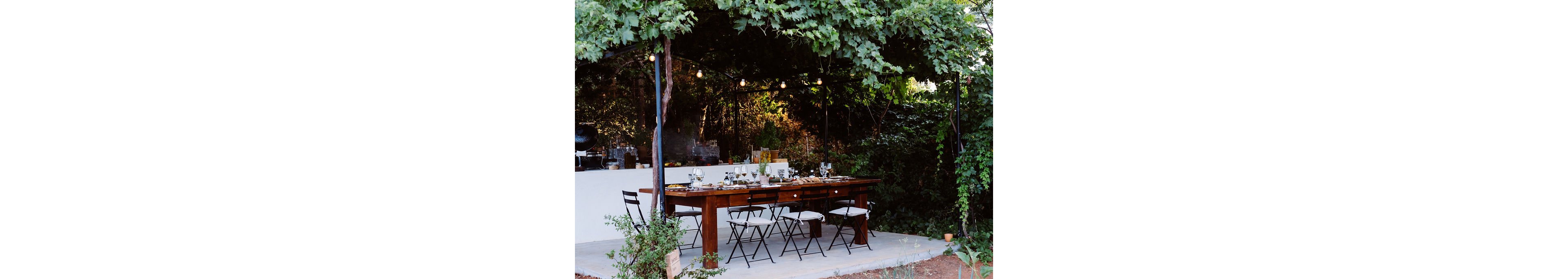 A long wooden dining table is set outdoors beneath a lush green canopy of vines and hanging string lights. Black metal chairs with white cushions surround the table, which is arranged with glassware and table settings for a meal. In the background, a person stands at a white outdoor bar or kitchen area, partially shaded by the foliage. The scene suggests an intimate garden gathering or al fresco dinner in a tranquil, natural setting.