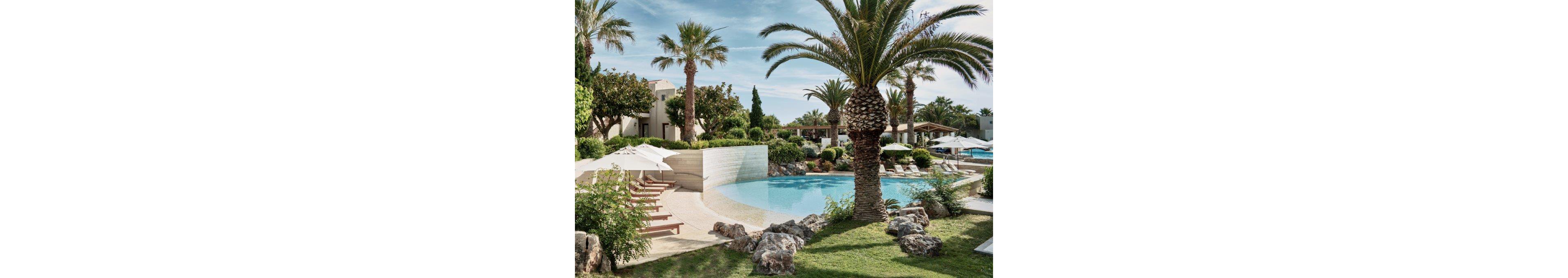A serene resort swimming pool is surrounded by palm trees, lush greenery, and landscaped rocks. Sun loungers with white umbrellas line the curved poolside, creating an inviting relaxation area. In the background, low-rise resort buildings and additional seating areas are visible under a clear sky. The overall scene suggests a peaceful vacation setting in a warm, tropical climate.