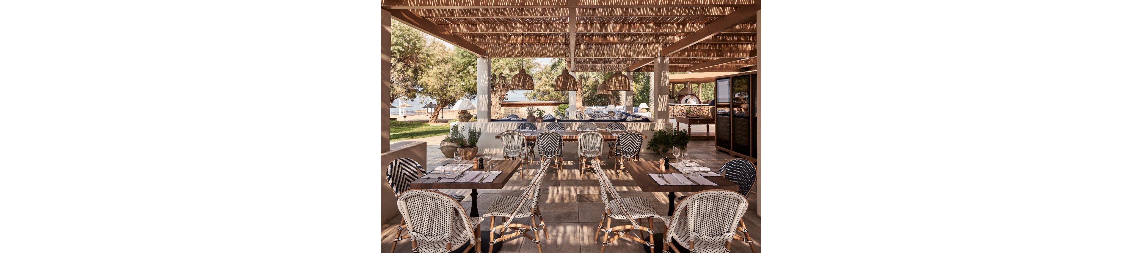 Stylish outdoor restaurant seating is arranged beneath a woven pergola roof. Wooden tables are set with placemats, glassware, and cutlery, surrounded by white and patterned wicker chairs. Natural tones, rattan light fixtures, and potted plants create a warm, rustic Mediterranean feel. In the background, trees and glimpses of the sea suggest a relaxed coastal dining experience.
