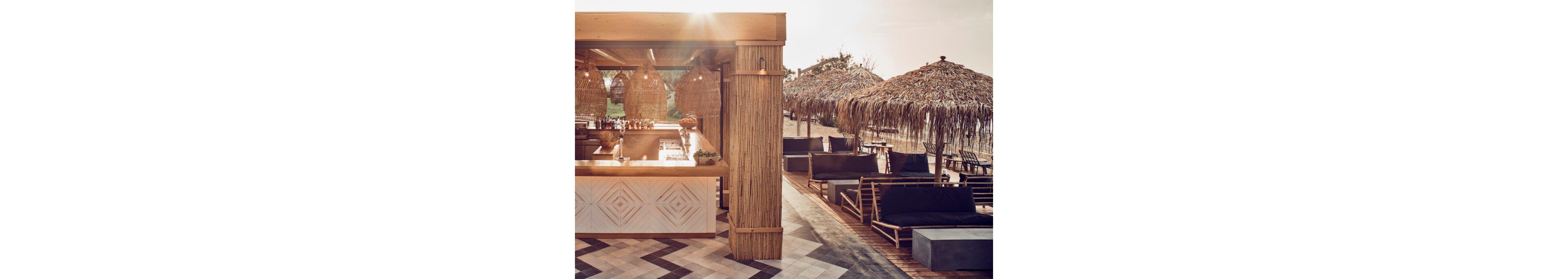 A contemporary open-air bar stands beside a sandy beach, featuring a wooden counter and woven pendant lights. Lounge chairs with dark cushions are arranged under thatched umbrellas facing the shoreline. The scene is bathed in warm, late-afternoon light, creating a relaxed vacation atmosphere. Natural materials and neutral tones emphasize a coastal, bohemian design style.