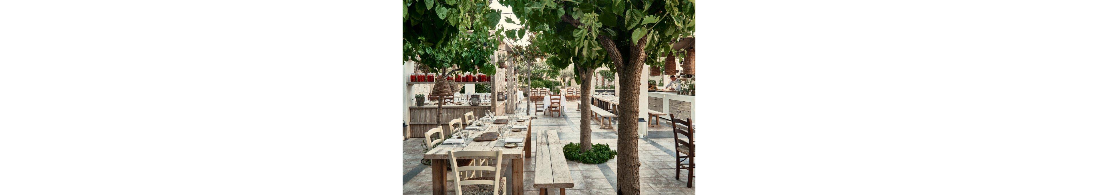 A rustic outdoor dining area is set with long wooden tables and simple chairs beneath leafy trees. Glassware, plates, and cutlery are neatly arranged, suggesting preparation for a meal service. The terrace features stone flooring, white walls, and natural wood accents that create a Mediterranean courtyard feel. In the background, additional seating and a partially visible open kitchen add depth and a relaxed restaurant atmosphere.