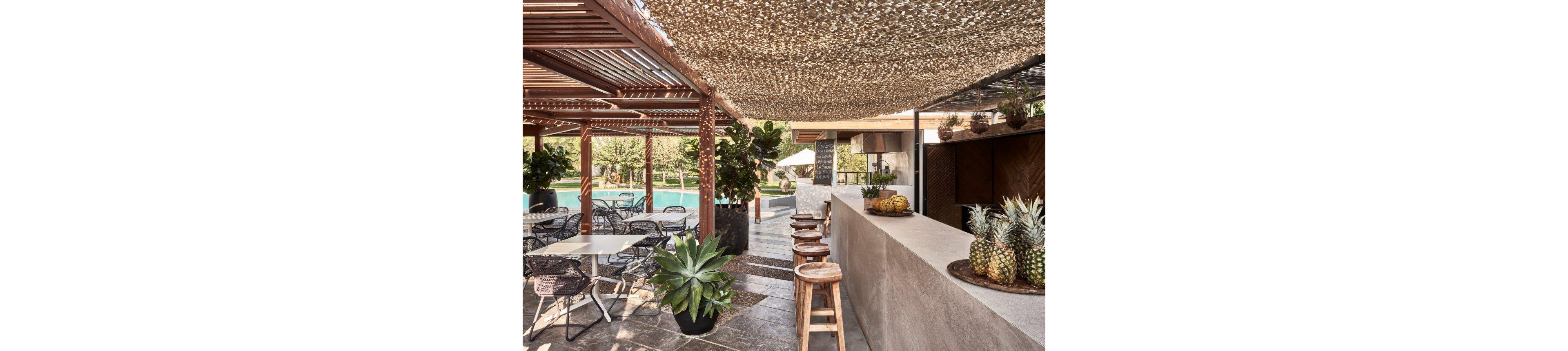 An open-air bar is set beside a swimming pool, shaded by a patterned canopy and wooden pergola. A bartender stands behind a long concrete counter topped with pineapples and decorative plants. Bar stools line the counter while metal chairs and tables provide seating in the foreground. Lush greenery and trees surround the area, creating a relaxed resort atmosphere.