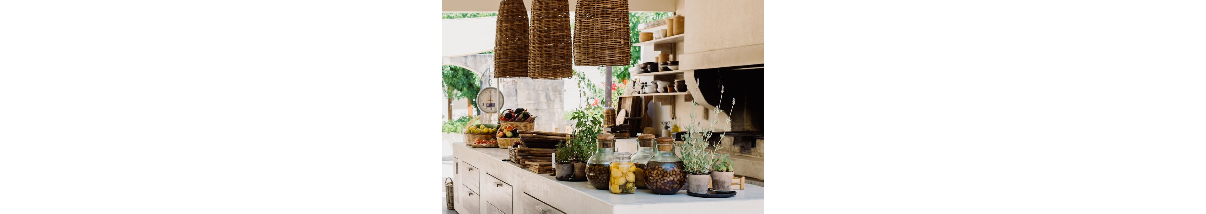 A bright rustic outdoor kitchen features a long white counter topped with jars, herbs, and fresh produce. Large woven wicker pendant lights hang from the ceiling, creating a warm focal point above the workspace. Open shelves hold dishes and kitchen items, while a built-in oven or grill is set into the wall. Natural wood textures and greenery contribute to a relaxed, Mediterranean-inspired atmosphere.