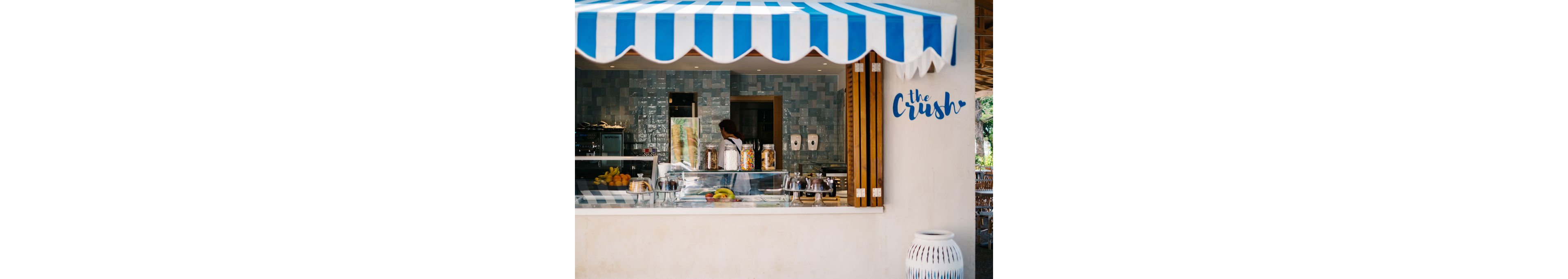 An open-air juice or snack bar features a blue and white striped awning above a serving counter. Inside, a worker stands behind glass displays filled with fresh fruit and jars of ingredients. The tiled interior wall and stainless-steel equipment create a clean, modern look. On the exterior wall, the text "the Crush" is visible, adding a playful touch to the scene.