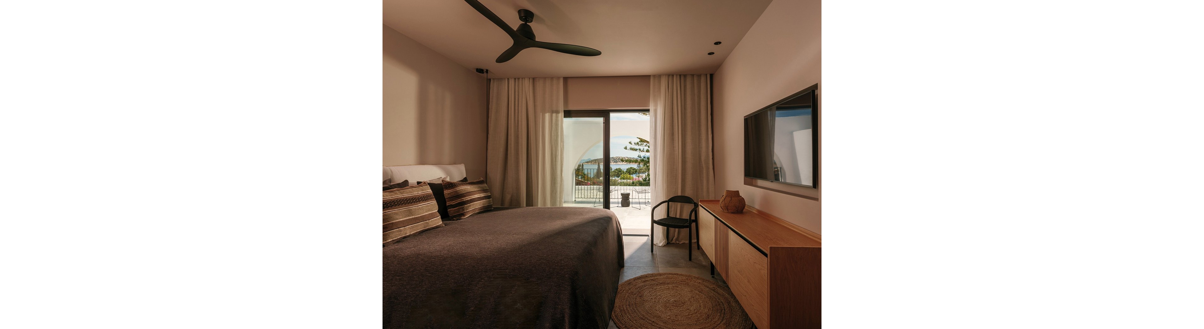 A contemporary bedroom features a large bed with striped pillows and neutral bedding. Floor-to-ceiling curtains frame a glass door that opens onto a balcony overlooking the sea and distant hills. A sleek ceiling fan, wall-mounted television, and minimalist wooden console with a decorative vase enhance the modern design. Warm, earthy tones and soft lighting create a calm, inviting atmosphere.
