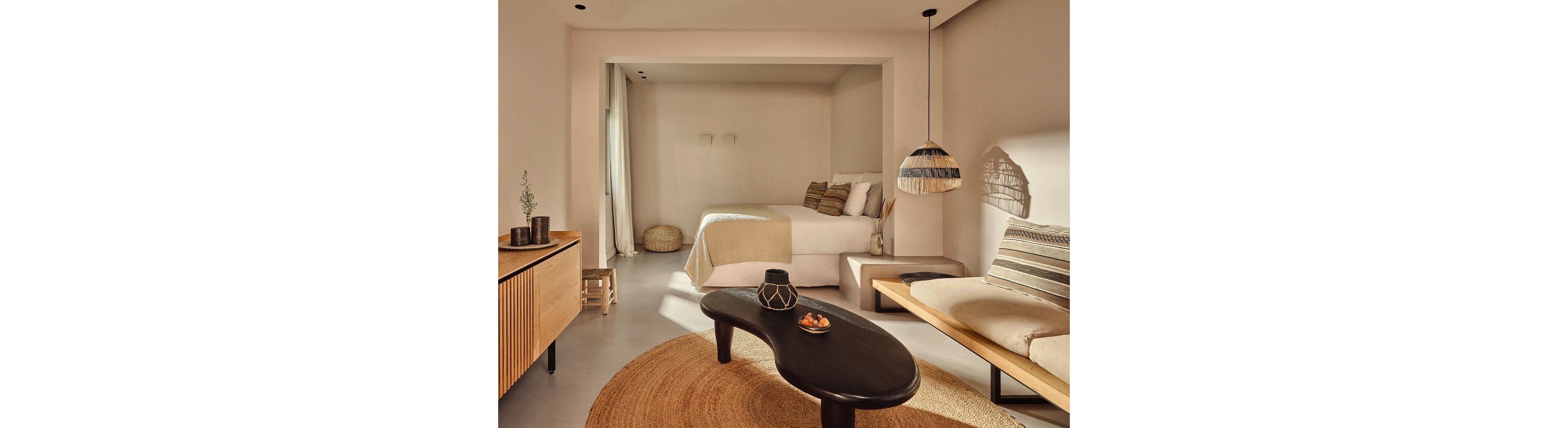 Warm natural light fills a minimalist bedroom and lounge area decorated in soft neutral tones. A bed with beige linens sits recessed in an alcove, complemented by woven baskets and simple wall lights. In the foreground, a dark organic-shaped coffee table stands on a round jute rug beside a low wooden bench with striped cushions. A wooden sideboard, hanging woven lamp, and subtle decorative objects enhance the cozy, contemporary atmosphere.