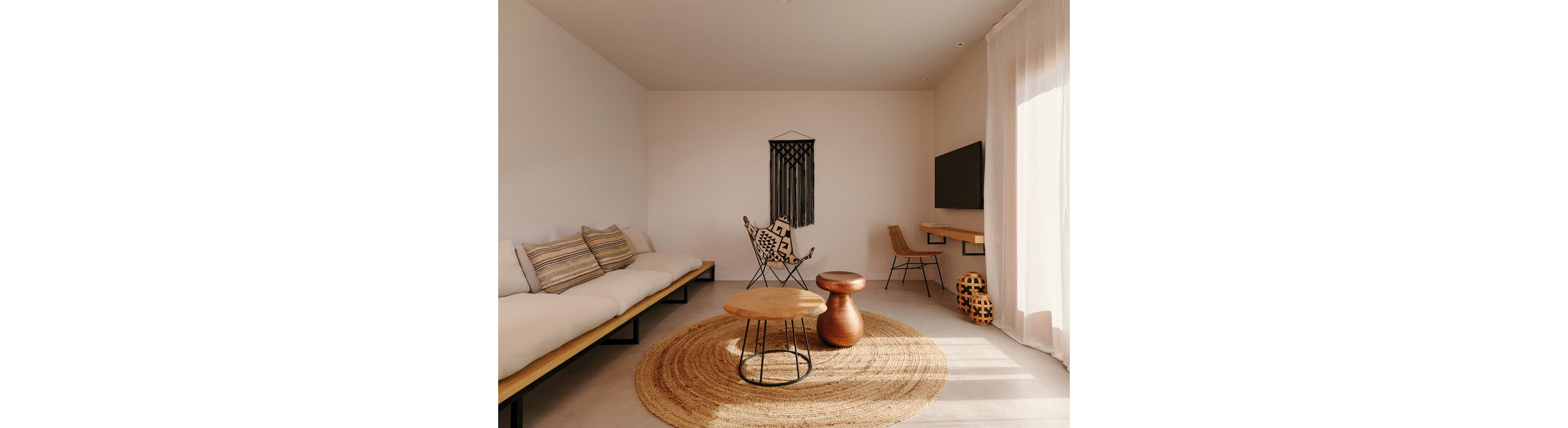 A minimalist living room features a long low sofa with striped cushions along one wall. In the center, round wooden coffee tables sit on a large woven jute rug, paired with a metal-framed accent chair. A wall-mounted television and simple desk with a chair occupy the opposite side, complemented by a patterned basket on the floor. Soft daylight filters through sheer curtains, highlighting the warm neutral tones and bohemian decor elements.