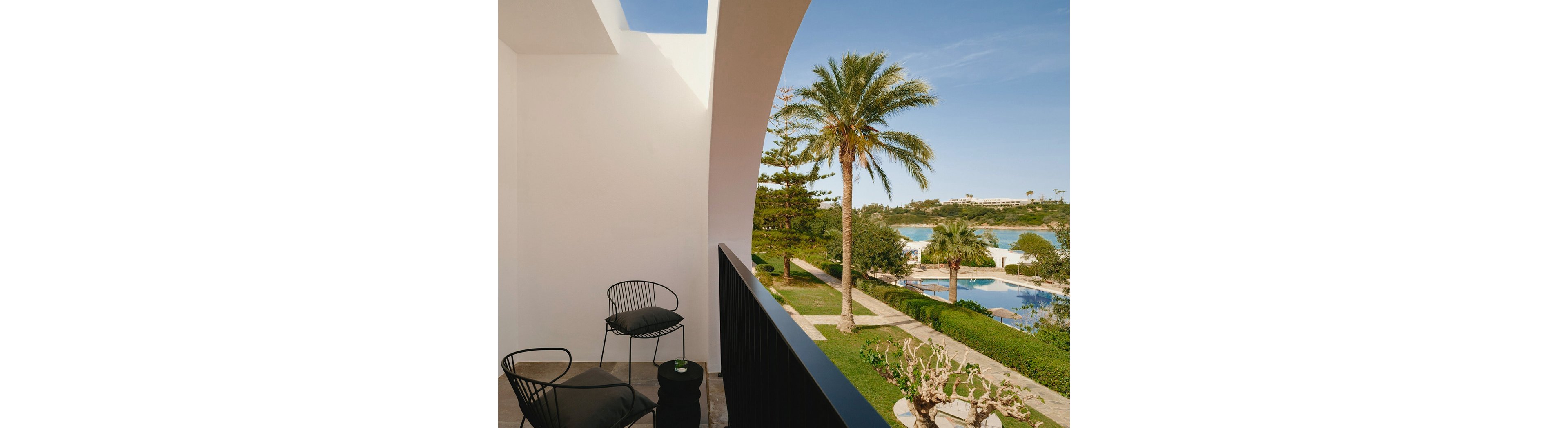 A minimalist balcony with two black chairs and a small side table looks out over a landscaped resort garden. Palm trees and manicured hedges frame a bright blue swimming pool below. Beyond the pool, a calm bay and distant buildings sit under a clear blue sky. The scene feels relaxed and inviting, ideal for vacation or leisure imagery.