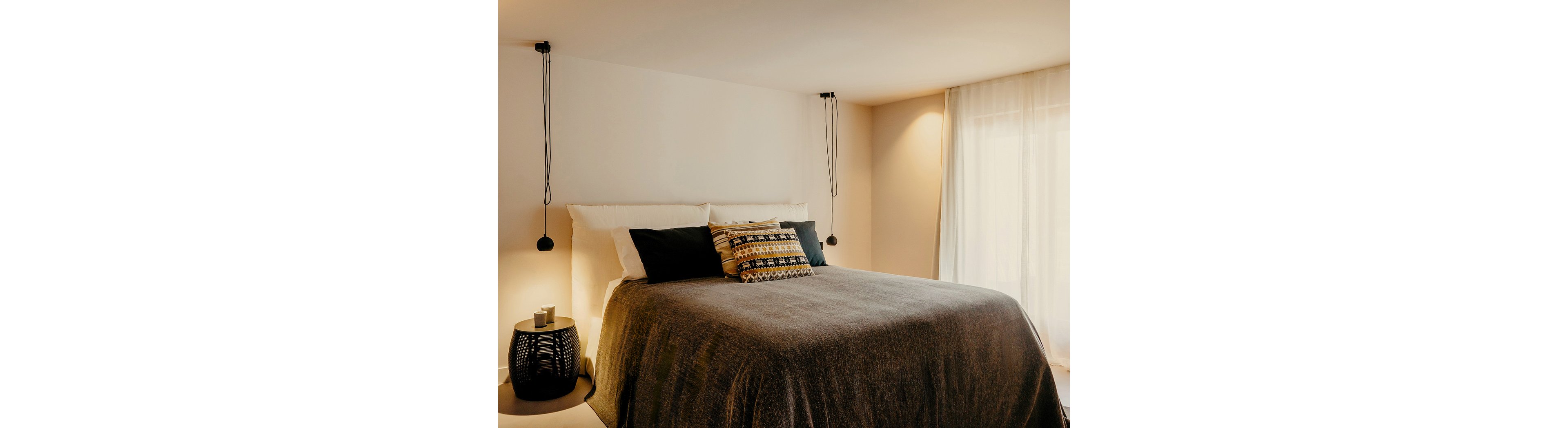 A neatly made double bed with layered pillows and a textured blanket is the focal point of a modern bedroom. Warm ambient light filters through sheer curtains on the right, creating a soft and inviting atmosphere. Minimalist pendant lights hang on either side of the bed, and a small side table with a candle adds a decorative touch. The overall scene feels calm, contemporary, and comfortable.