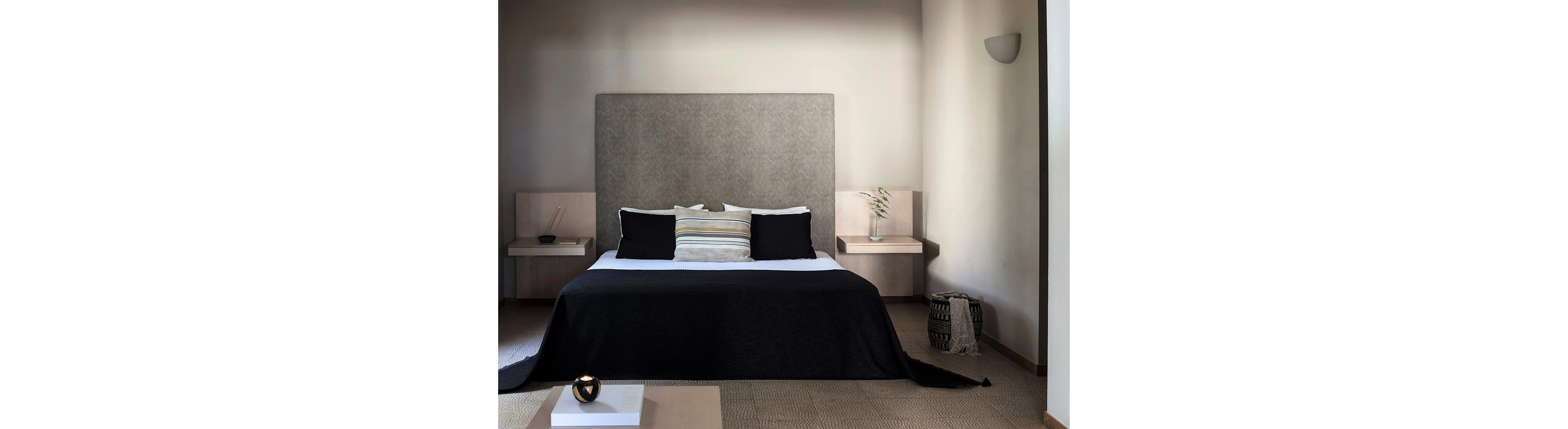 A contemporary bedroom features a large bed with a tall upholstered headboard and a dark bedspread. Neutral-colored pillows add subtle contrast against the soft beige walls. Floating bedside tables hold simple decor, including a vase with greenery and small bowls. A woven basket and a low table with a decorative object complete the minimalist, serene setting.