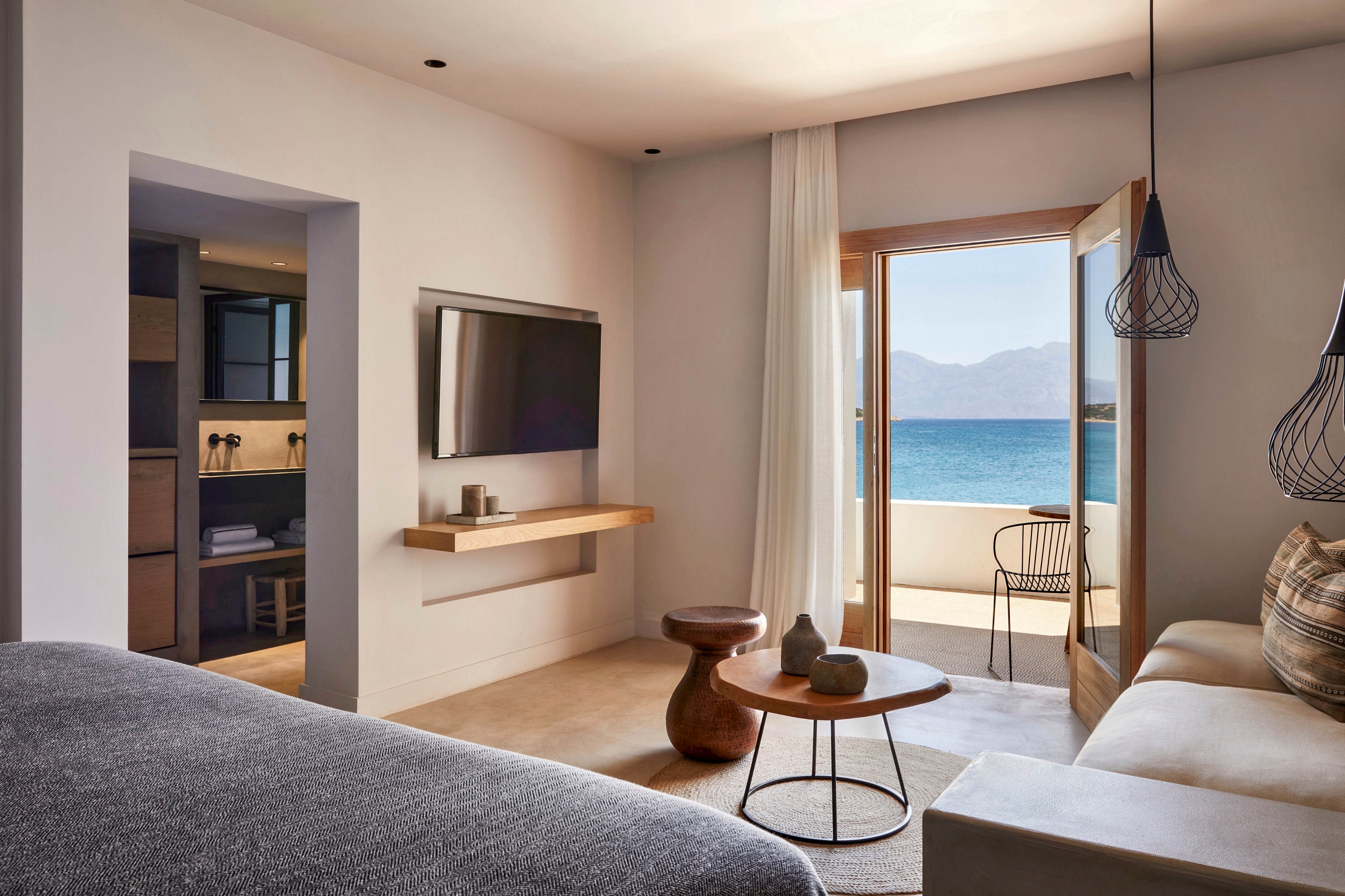 A contemporary hotel room featuring a minimalist design with a wall-mounted TV, wooden furniture, and a cozy seating area. The room opens to a balcony with a stunning ocean view, framed by large glass doors. Neutral tones and natural light create a serene and inviting atmosphere.