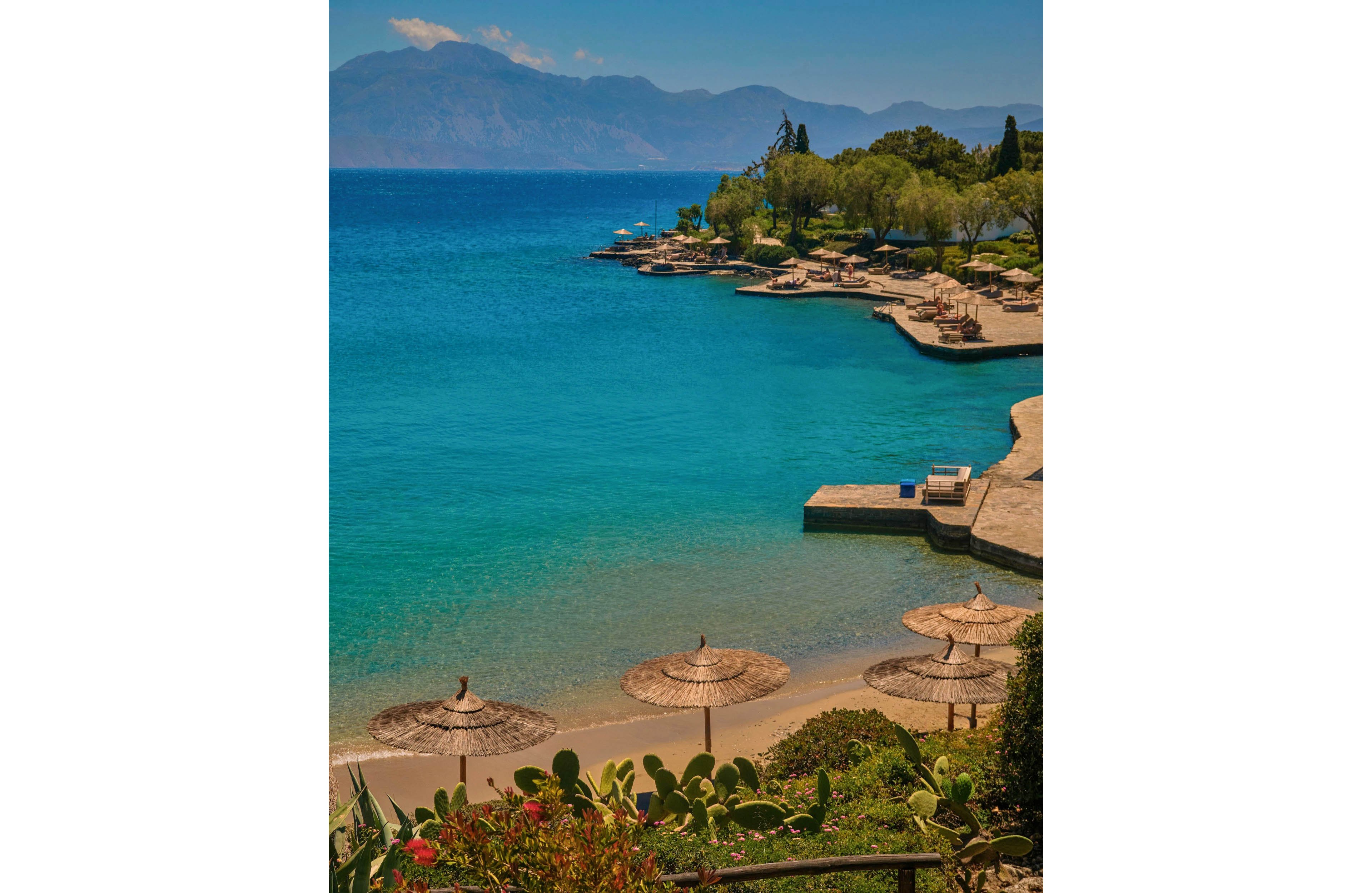 A tranquil seaside resort features clear turquoise water gently meeting a sandy beach. Straw parasols and sun loungers line the shore and stone terraces, inviting visitors to relax by the sea. Lush greenery and cactus plants frame the foreground, while distant mountains rise across the bay under a warm, hazy sky. The scene evokes a peaceful summer holiday atmosphere on a calm coastal shoreline.