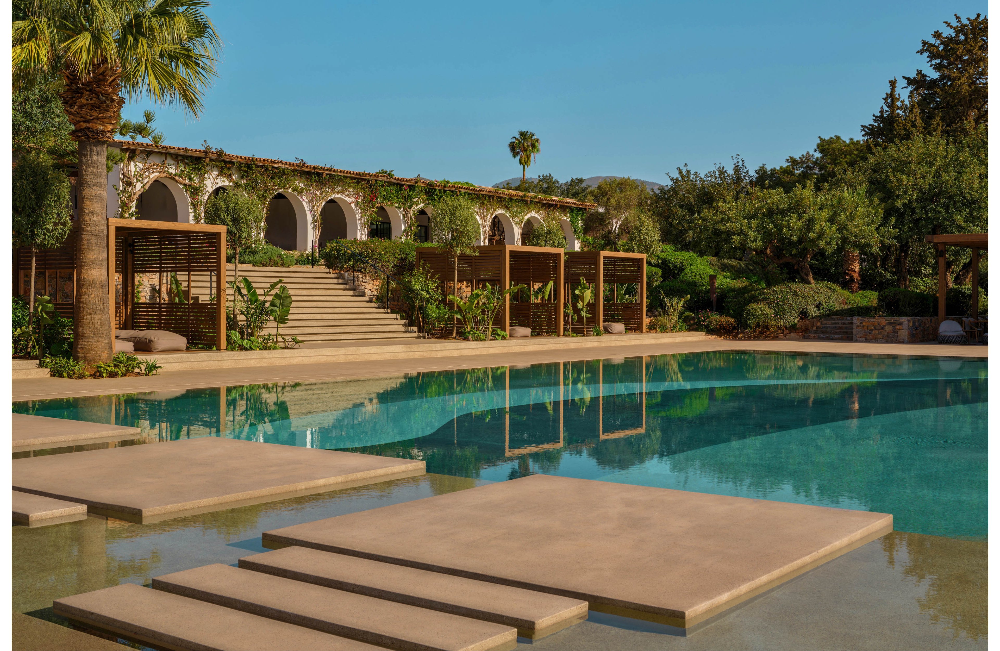 A serene outdoor swimming pool sits in front of a Mediterranean-style building with stone arches and lush greenery. Wide steps lead up to a terrace framed by pergolas and tropical plants. Palm trees and dense foliage surround the area, creating a secluded resort atmosphere. The calm water reflects the architecture and garden, emphasizing relaxation and leisure.