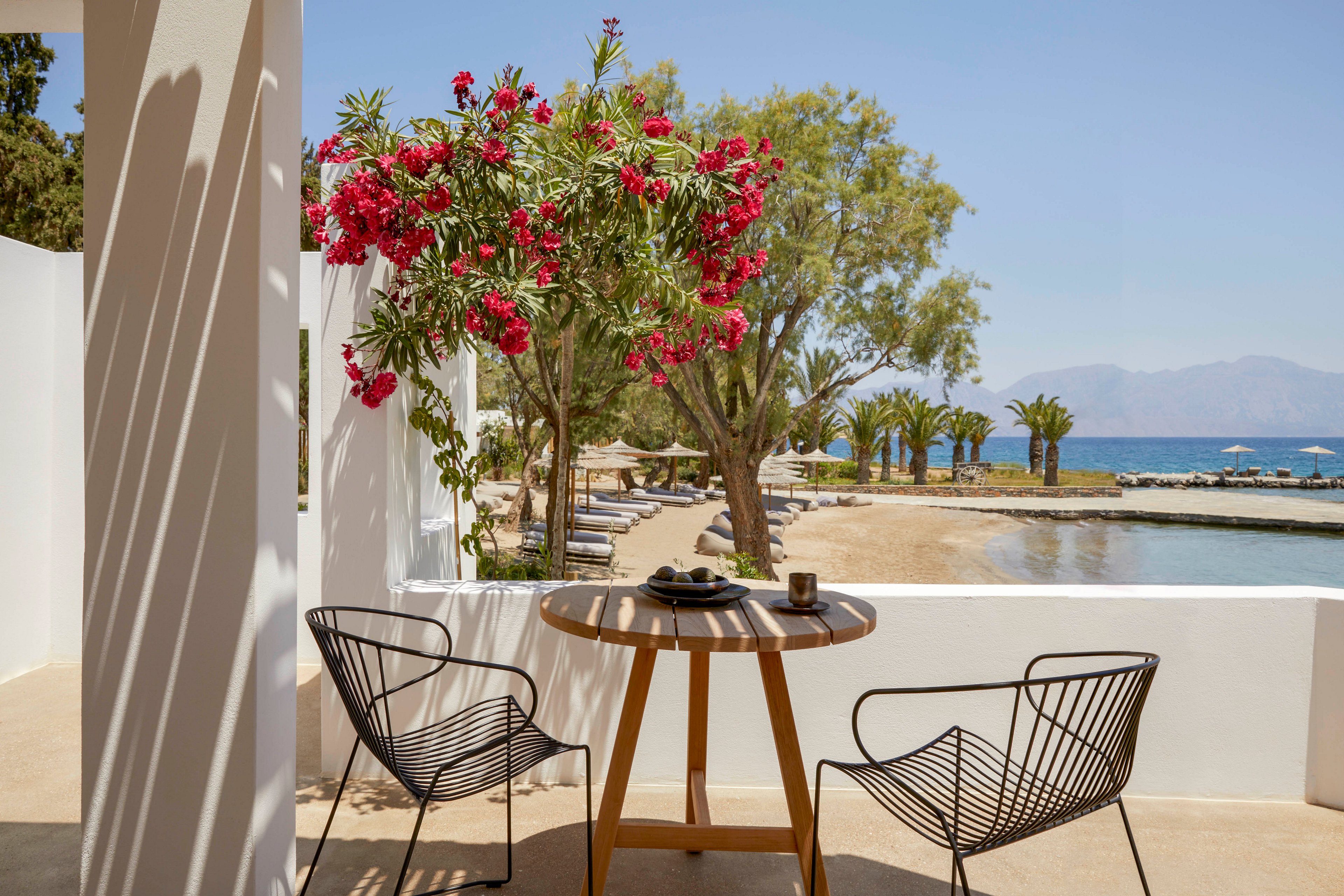 A modern terrace features a round wooden table and two black metal chairs overlooking a sandy beach. Bright pink flowers and lush green trees frame the view toward the calm blue sea. Sun loungers and umbrellas line the shoreline under a clear sky, suggesting a relaxing vacation setting. Strong sunlight creates clean shadows and a warm, inviting atmosphere.