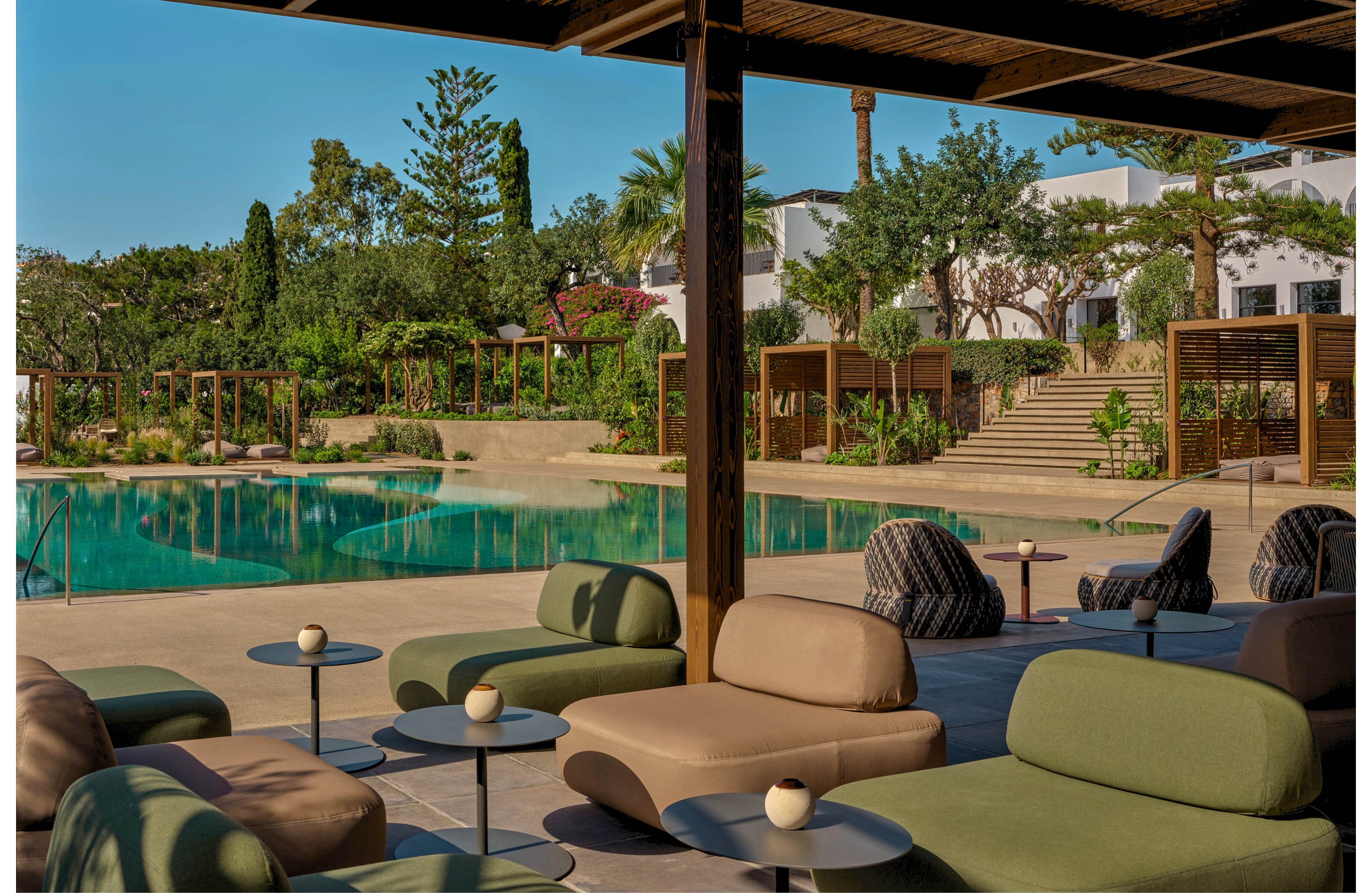 A contemporary open-air lounge area features cushioned chairs and low tables arranged under a wooden pergola. Beyond the seating, a clear turquoise swimming pool is surrounded by lush greenery and manicured landscaping. White resort buildings and palm trees rise in the background, creating a relaxed vacation atmosphere. The scene is bathed in warm daylight, emphasizing the modern, minimalist design and tranquil setting.