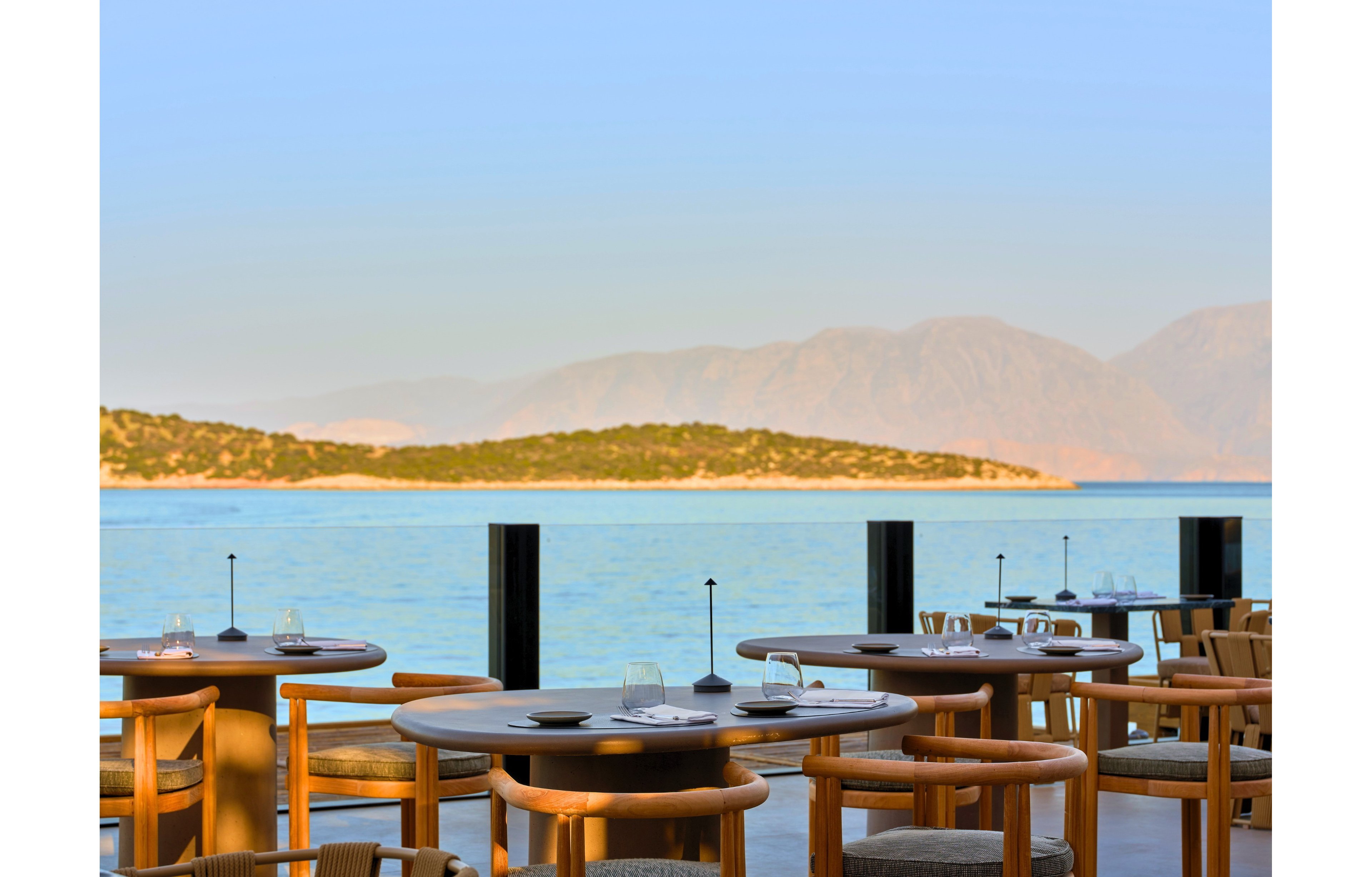An open-air restaurant terrace overlooks a calm blue sea with distant mountains under a clear sky. Round wooden tables and chairs are neatly arranged, some set with glassware and plates. The scene suggests a peaceful dining experience by the water, with soft natural light enhancing the coastal atmosphere. No people are present, emphasizing the tranquil and inviting setting.