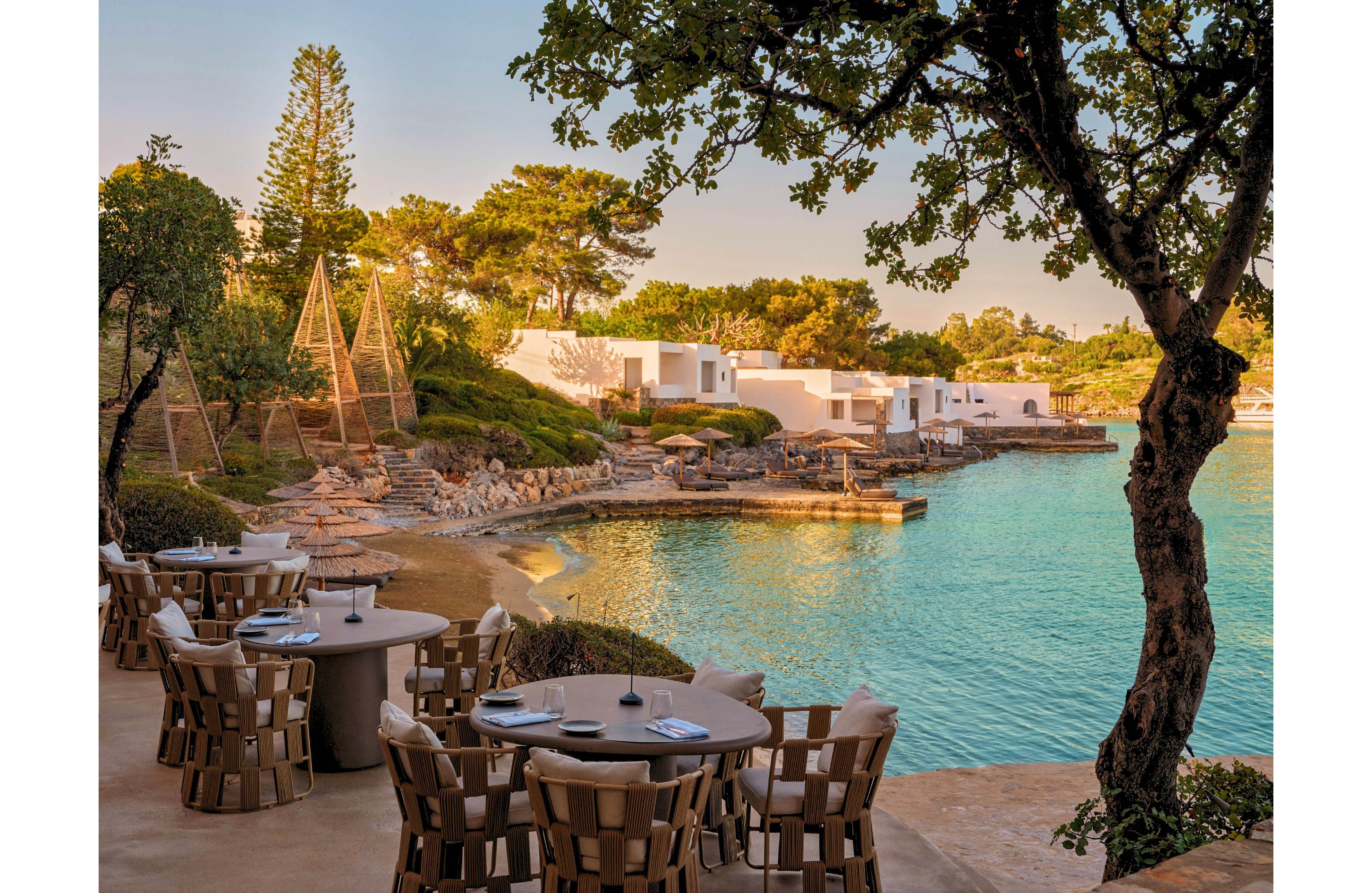 An elegant outdoor restaurant overlooks a calm turquoise lagoon bordered by stone terraces and modern white villas. Round tables with woven chairs are set with plates and napkins, ready for guests in the warm evening light. Tall sculptural wooden structures rise among landscaped greenery and trees along the shoreline. The overall scene feels tranquil and luxurious, ideal for seaside dining and relaxation.