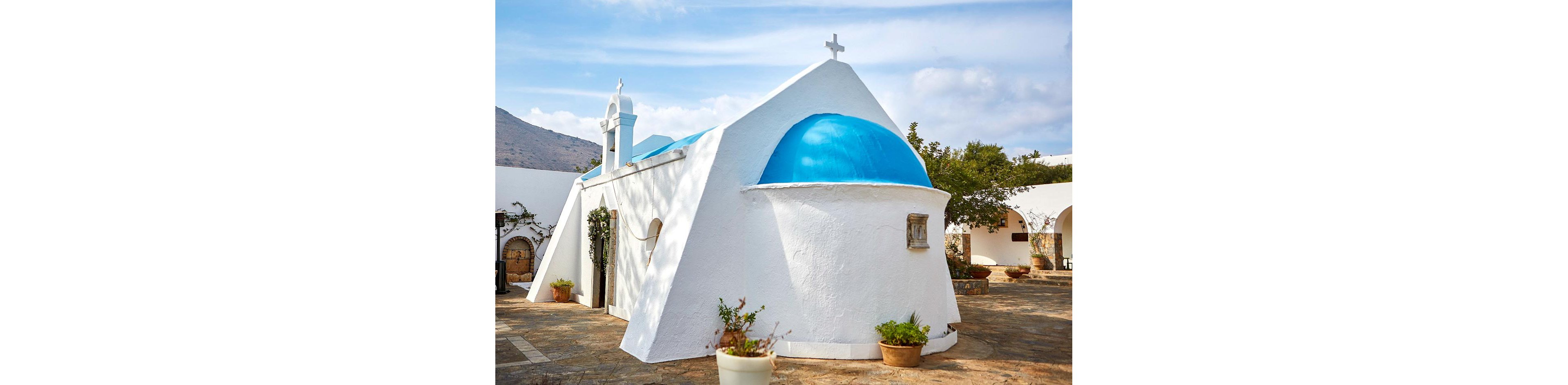 A small whitewashed chapel with a bright blue dome stands in a sunlit courtyard. The building features simple architectural lines, a bell tower, and a cross on top. Terracotta pots with plants and vines decorate the stone-paved courtyard around the chapel. In the background, trees and distant hills appear under a partly cloudy sky, suggesting a peaceful Mediterranean setting.
