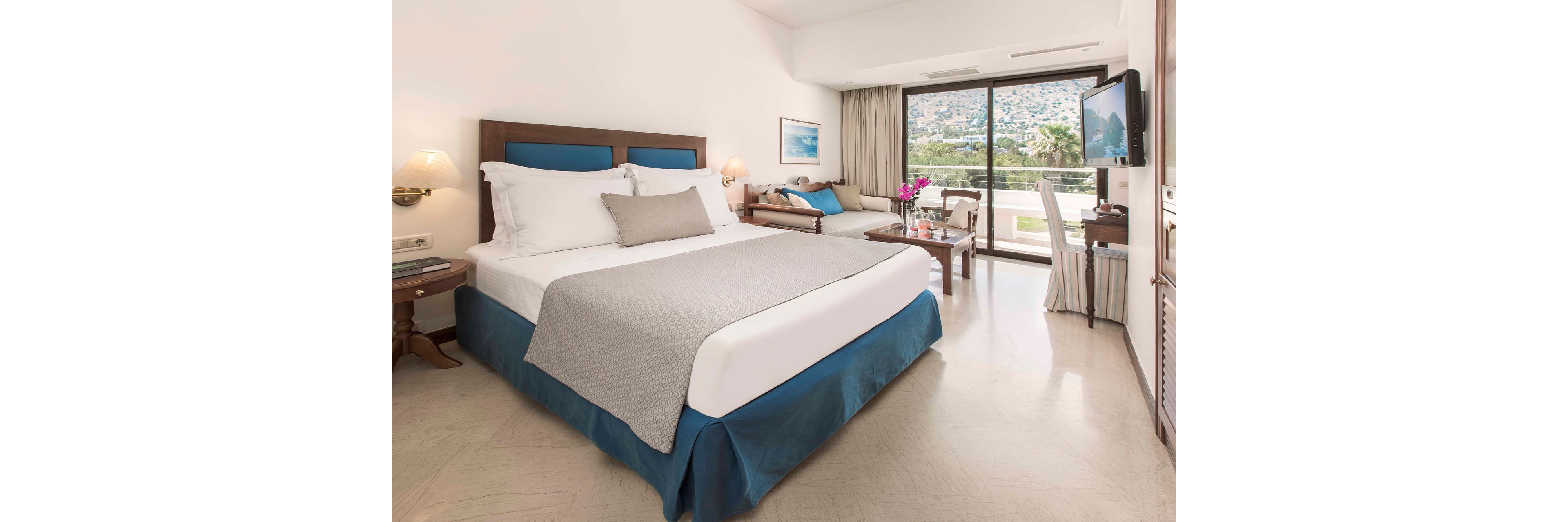 A neatly made double bed with white linens and a light gray blanket is the focal point of this bright hotel room. The space includes wooden bedside tables, a sofa seating area, and a small table with flowers. Large glass doors open to a balcony overlooking a scenic hillside and water. Soft neutral tones with blue accents create a calm, inviting atmosphere.