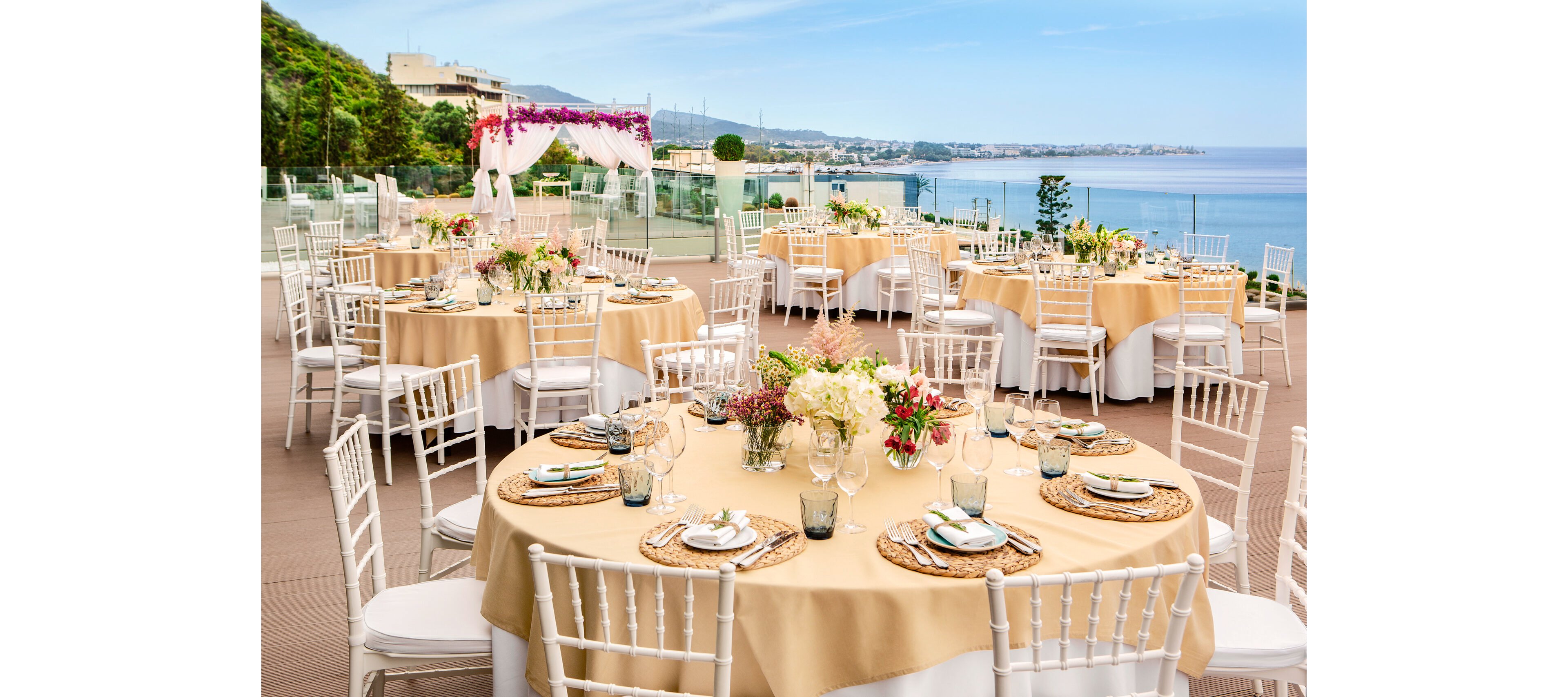 Round tables with beige tablecloths and white chairs are arranged on a terrace overlooking a coastal landscape. Floral centerpieces, glassware, and woven placemats decorate each table, suggesting a formal event or wedding reception. A flower-adorned archway stands near the edge of the terrace, framing the ocean and distant shoreline. The bright daylight, blue sky, and calm water create a cheerful, romantic atmosphere.