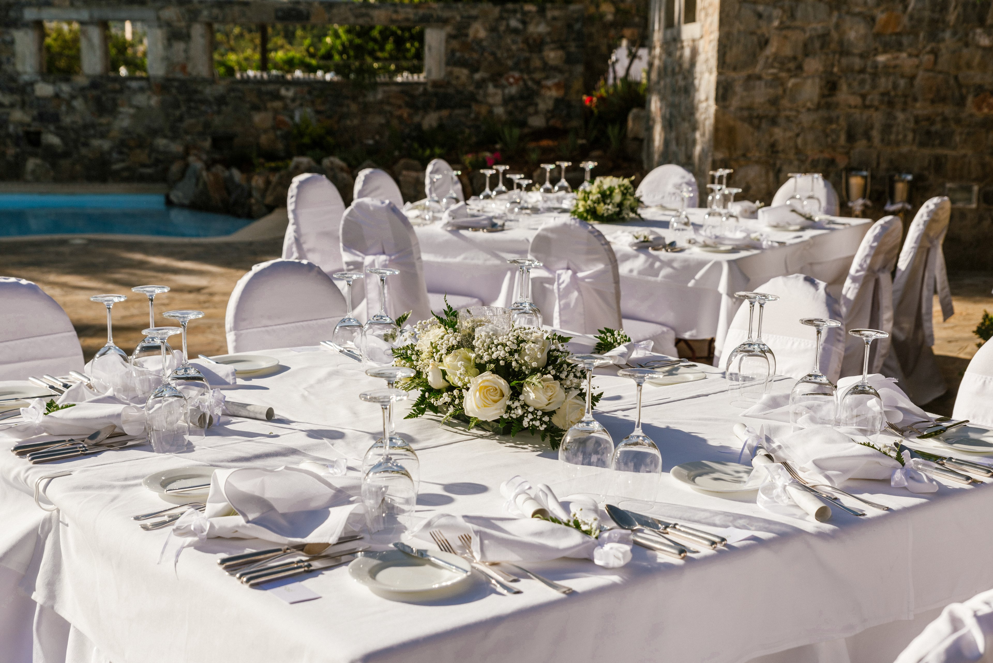 White-clothed tables are beautifully arranged for an outdoor reception in a sunny courtyard. Each place setting features neatly folded napkins, cutlery, plates, and multiple upside-down wine glasses. A floral centerpiece with white roses and greenery decorates the main table, adding a romantic touch. Chair covers and the stone wall backdrop create a formal, celebratory atmosphere near a small pool.