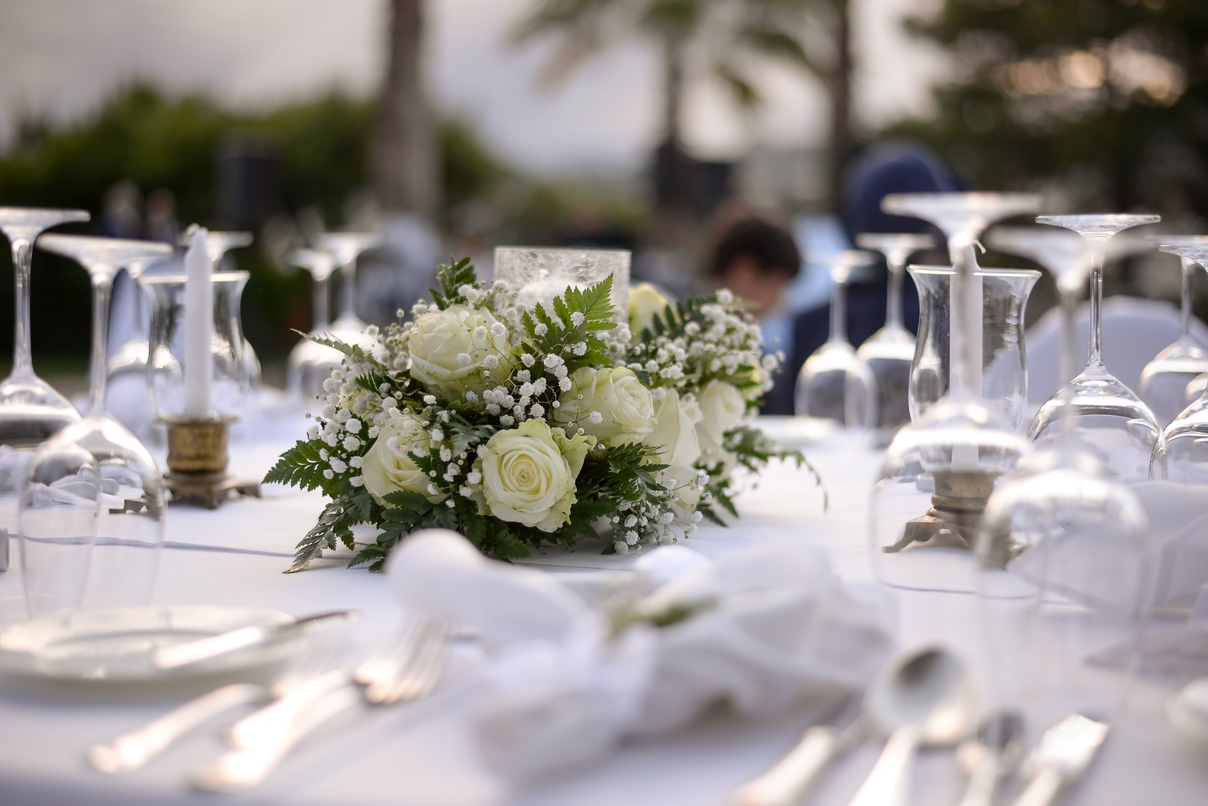 A beautifully set table features a floral centerpiece of white roses and baby's breath surrounding a glass candle holder. Polished glassware and cutlery are arranged neatly on a white tablecloth, suggesting a formal event or celebration. The soft, natural light and blurred background with trees and palm silhouettes create an outdoor, romantic atmosphere. The overall scene conveys sophistication and readiness for a special gathering or reception.