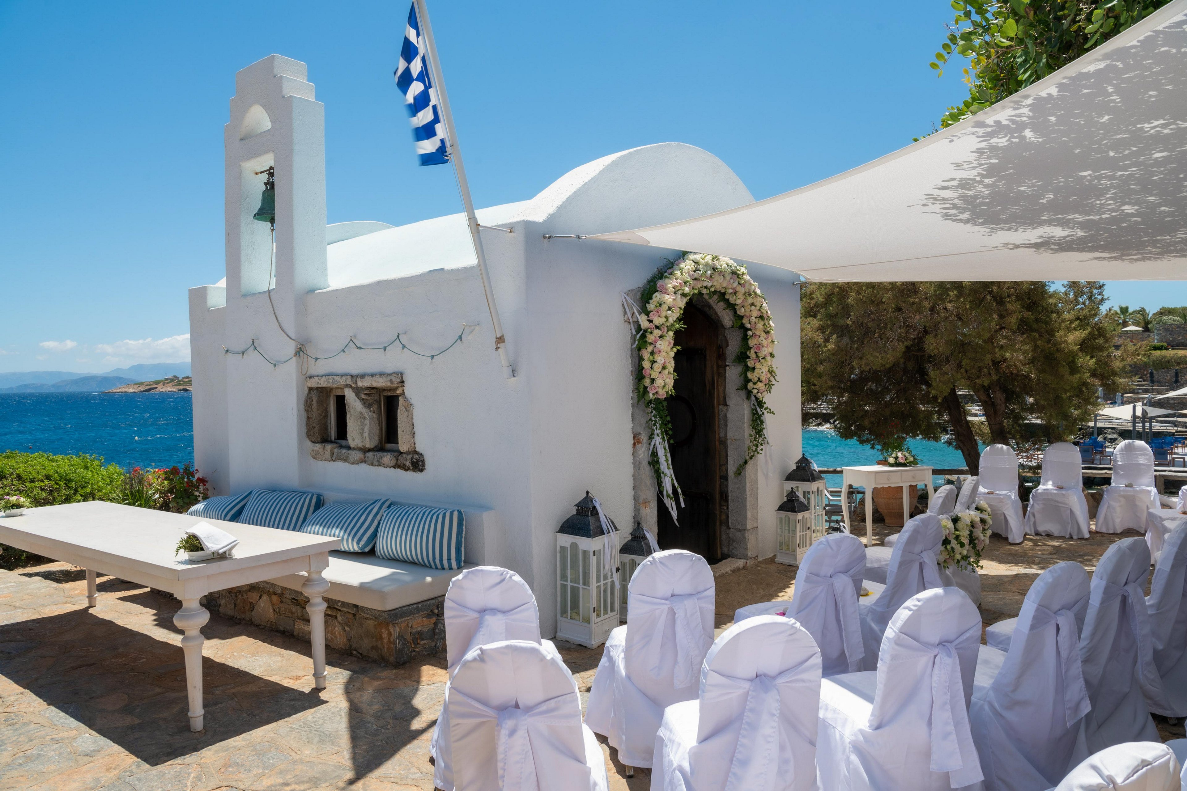 A small white chapel with a Greek flag stands beside a bright blue sea, decorated for a wedding ceremony. Rows of white-covered chairs face the entrance, which is adorned with floral garlands and greenery. A white table and lanterns enhance the elegant outdoor setting under a sunshade canopy. Trees and distant coastline complete the serene Mediterranean backdrop.