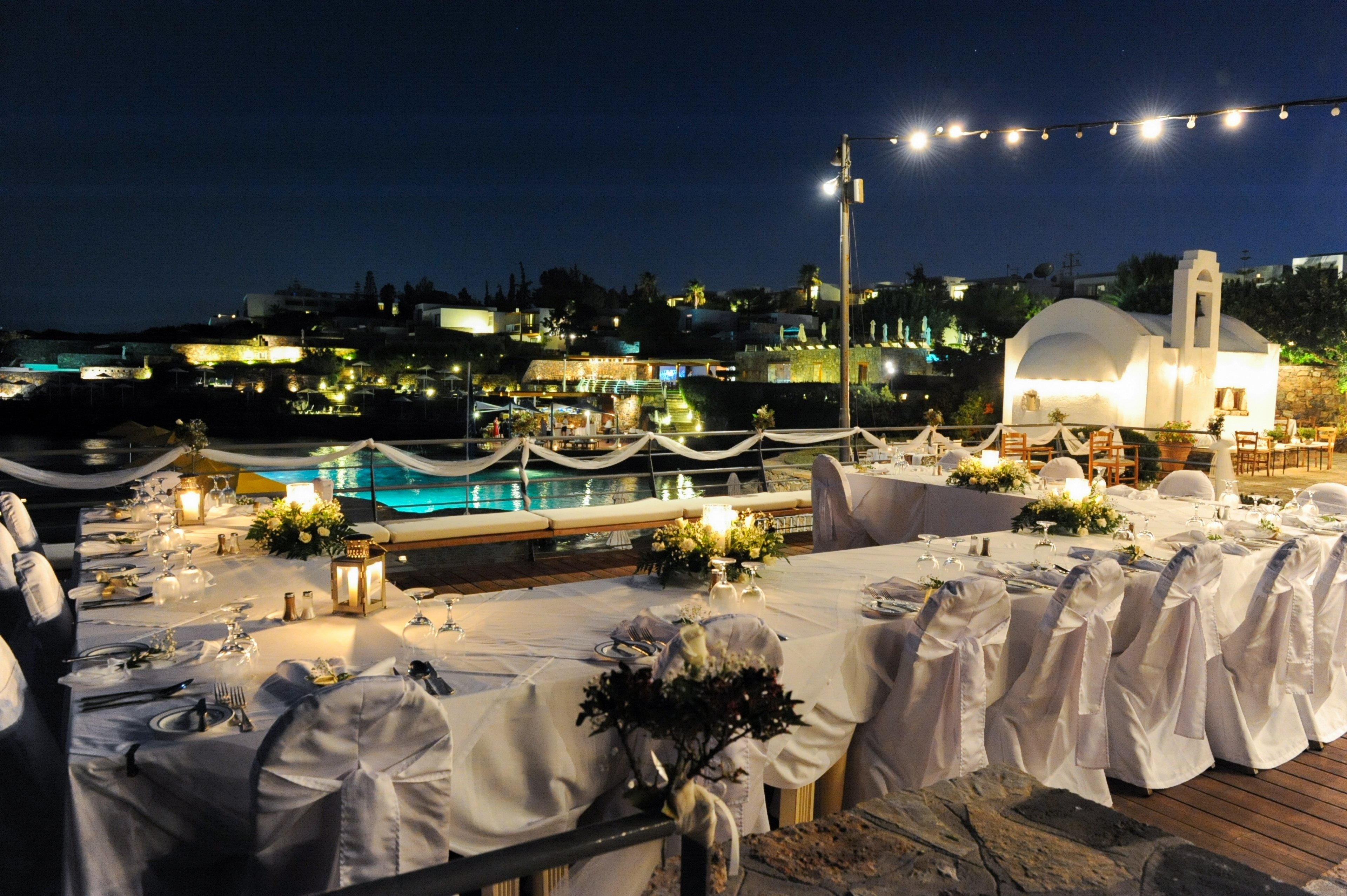 An elegantly arranged outdoor reception table is set beside a pool at night, with white chair covers and table linens creating a formal atmosphere. Soft lighting from string lights and lanterns illuminates the scene, highlighting floral centerpieces and neatly arranged place settings. In the background, buildings and a small white chapel-like structure are visible against the dark sky, adding a romantic coastal ambiance. The overall mood is festive and sophisticated, suitable for a wedding or special celebration.