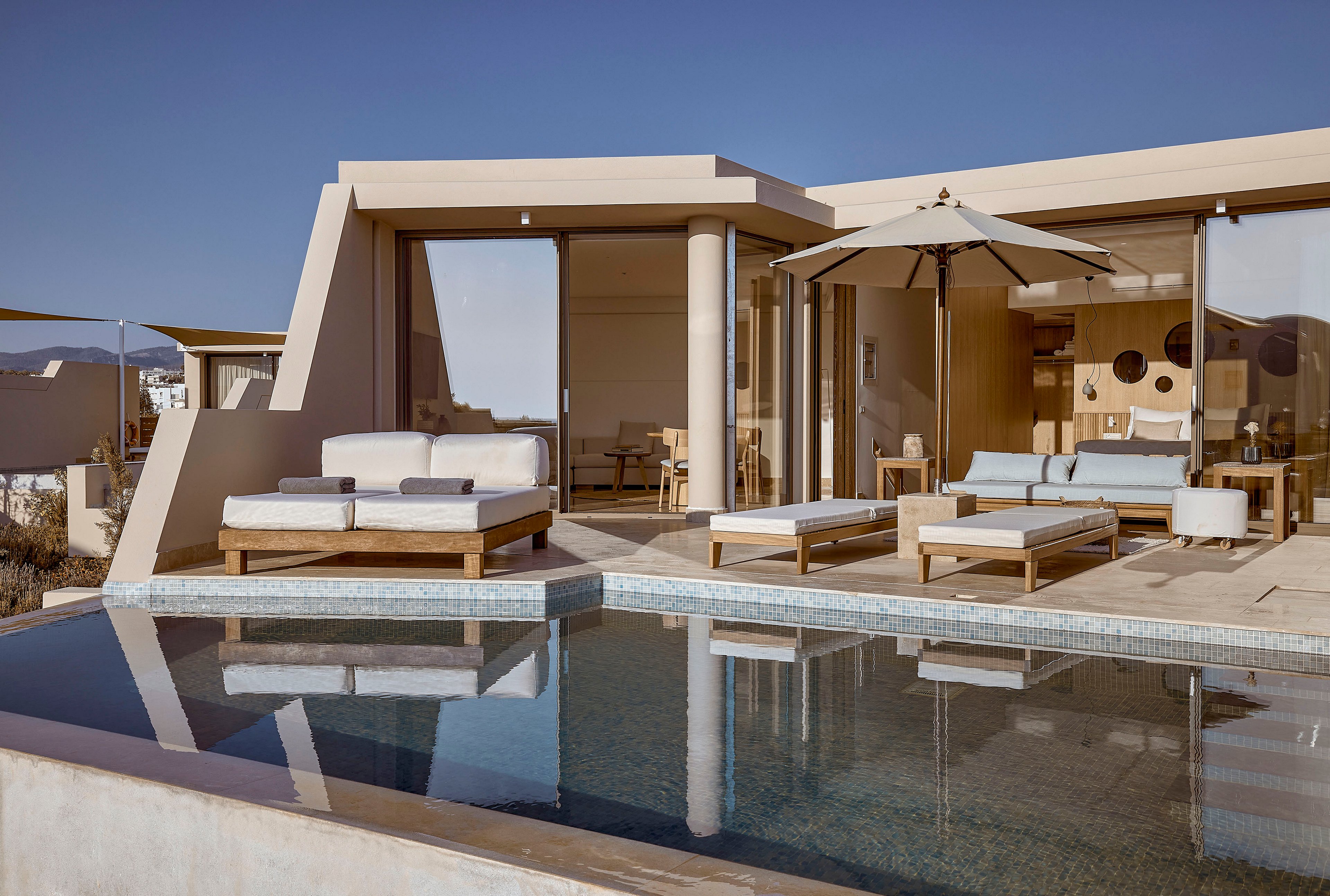 A contemporary villa features a private swimming pool in front of a spacious sun terrace. Minimalist lounge chairs and daybeds sit under a large patio umbrella, facing floor-to-ceiling glass doors. Warm neutral tones and clean architectural lines create a serene, upscale atmosphere. The scene is bathed in soft daylight, suggesting a relaxing vacation or resort setting.