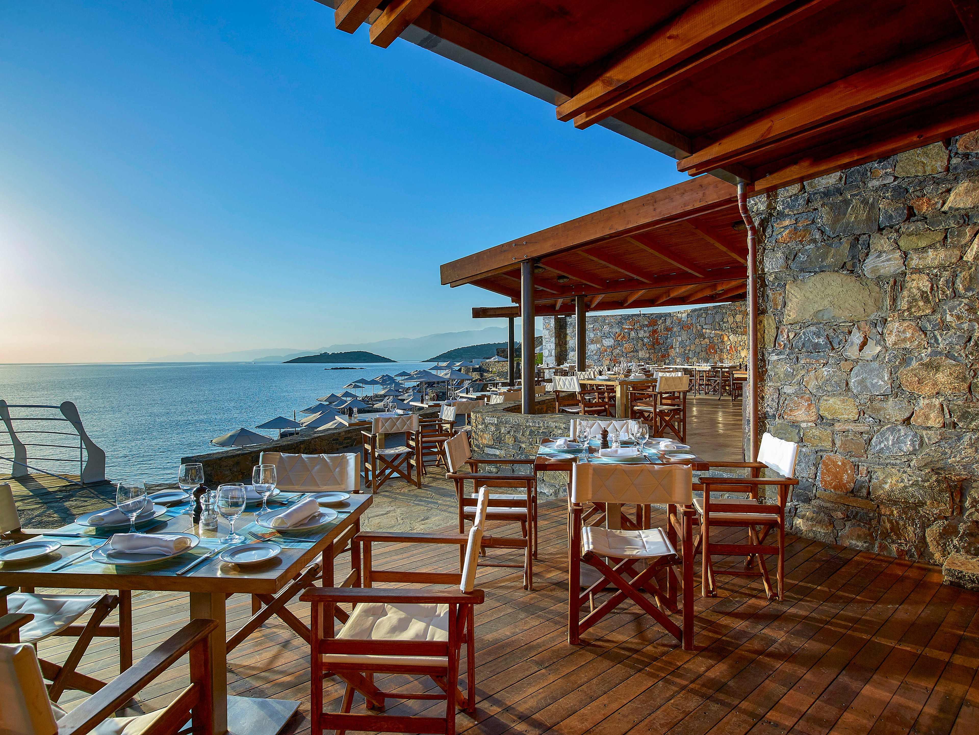 An open-air restaurant terrace overlooks a calm sea at sunset. Wooden tables and chairs are neatly arranged with glassware and plates ready for diners. Stone walls and a wooden roof structure frame the space, creating a rustic Mediterranean ambiance. In the background, lounge chairs line the waterfront and distant islands are visible on the horizon.