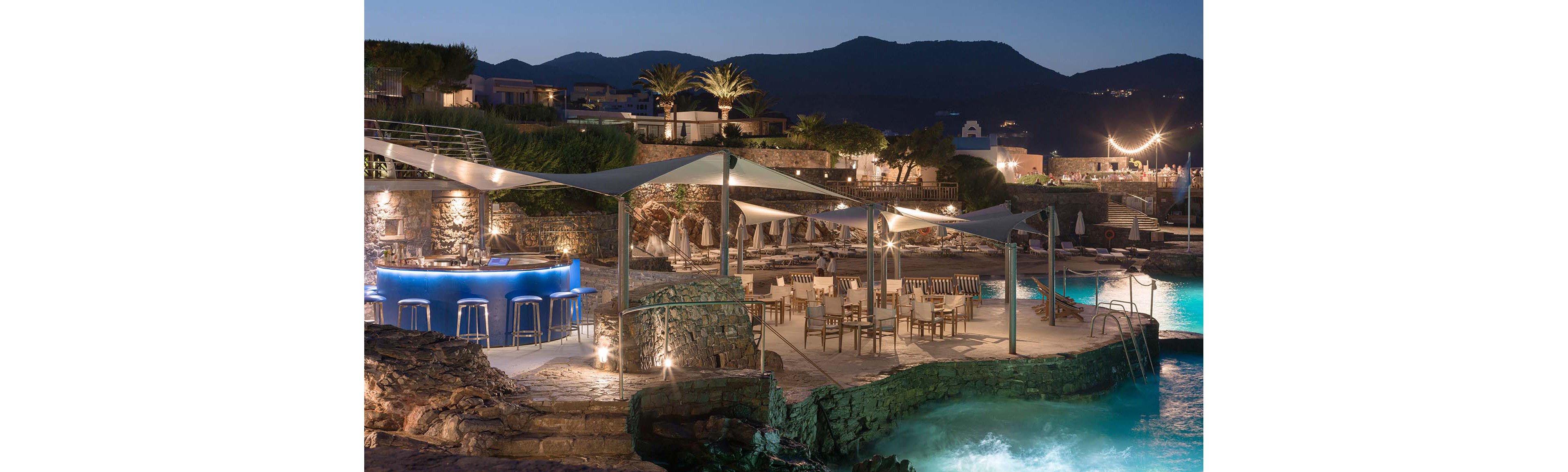 An upscale outdoor bar and dining area overlooks a turquoise seaside pool at twilight. Stone terraces, modern shade canopies, and neatly arranged tables and chairs create a sophisticated resort atmosphere. Warm ambient lighting highlights the bar counter, seating areas, and surrounding rock formations. In the background, palm trees and low buildings sit against a silhouette of distant mountains under a deep blue evening sky.