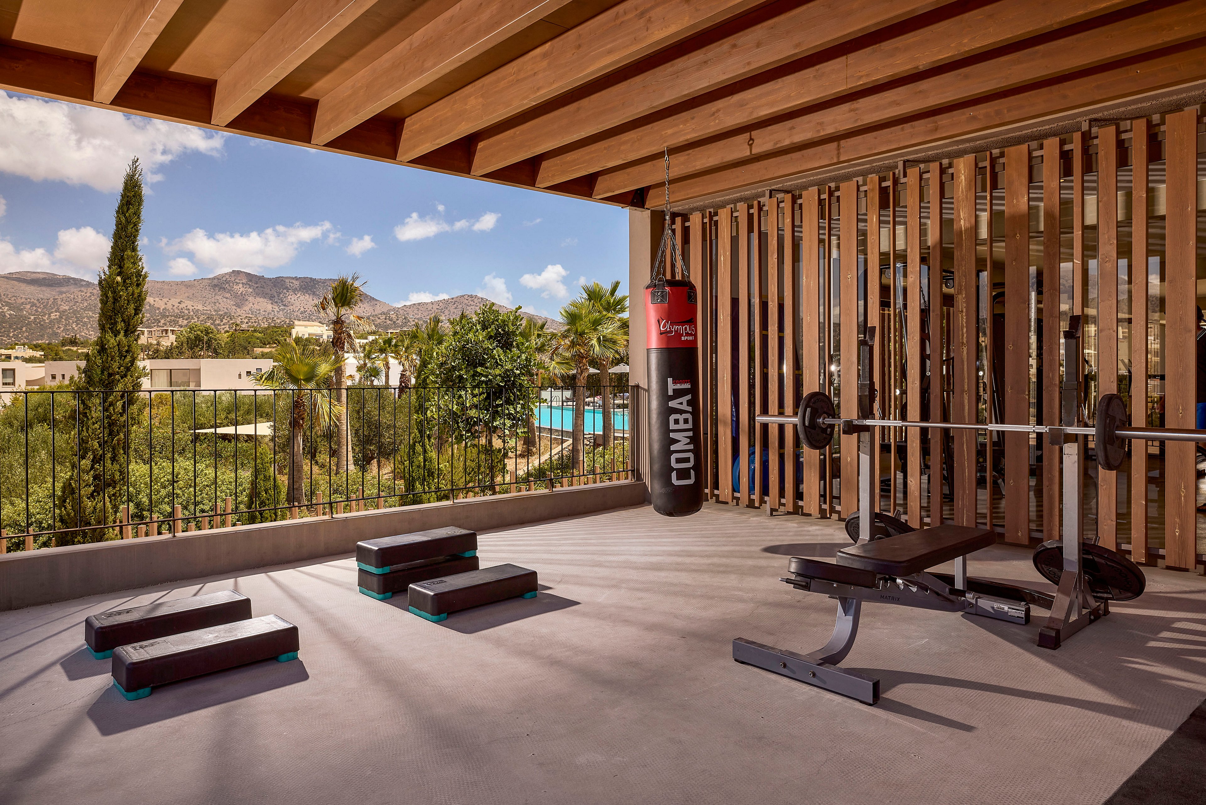 An open-air gym terrace features a hanging red punching bag labeled COMBAT alongside a weight bench and barbell. Several aerobic step platforms are arranged on the concrete floor under a wooden slatted roof. Vertical wooden beams frame views of palm trees, modern buildings, and distant mountains under a blue sky with scattered clouds. The scene suggests a tranquil yet energetic environment for fitness and combat training.