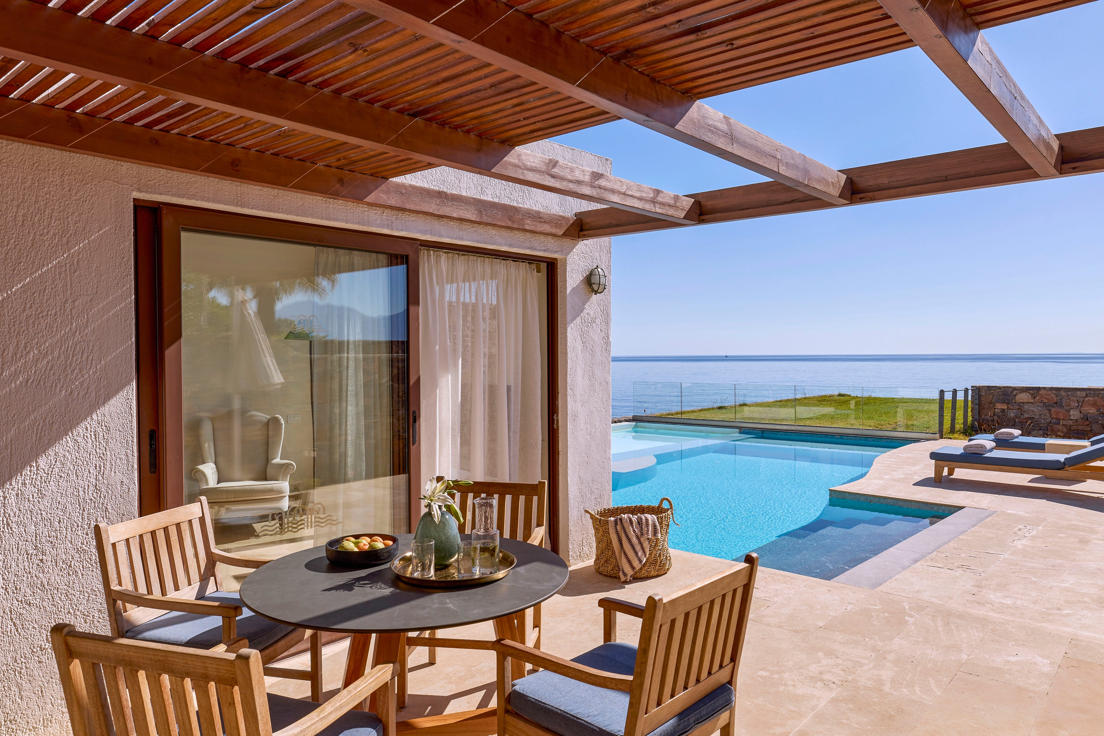Outdoor terrace of a modern coastal villa featuring a round dining table with wooden chairs under a slatted pergola. A sliding glass door with sheer curtains connects the interior to the sunlit patio. The scene includes a bright blue swimming pool, lounge chairs, and a view of the calm ocean beyond a glass railing. Decorative items like a fruit bowl, carafe, and woven bag add a relaxed vacation atmosphere.