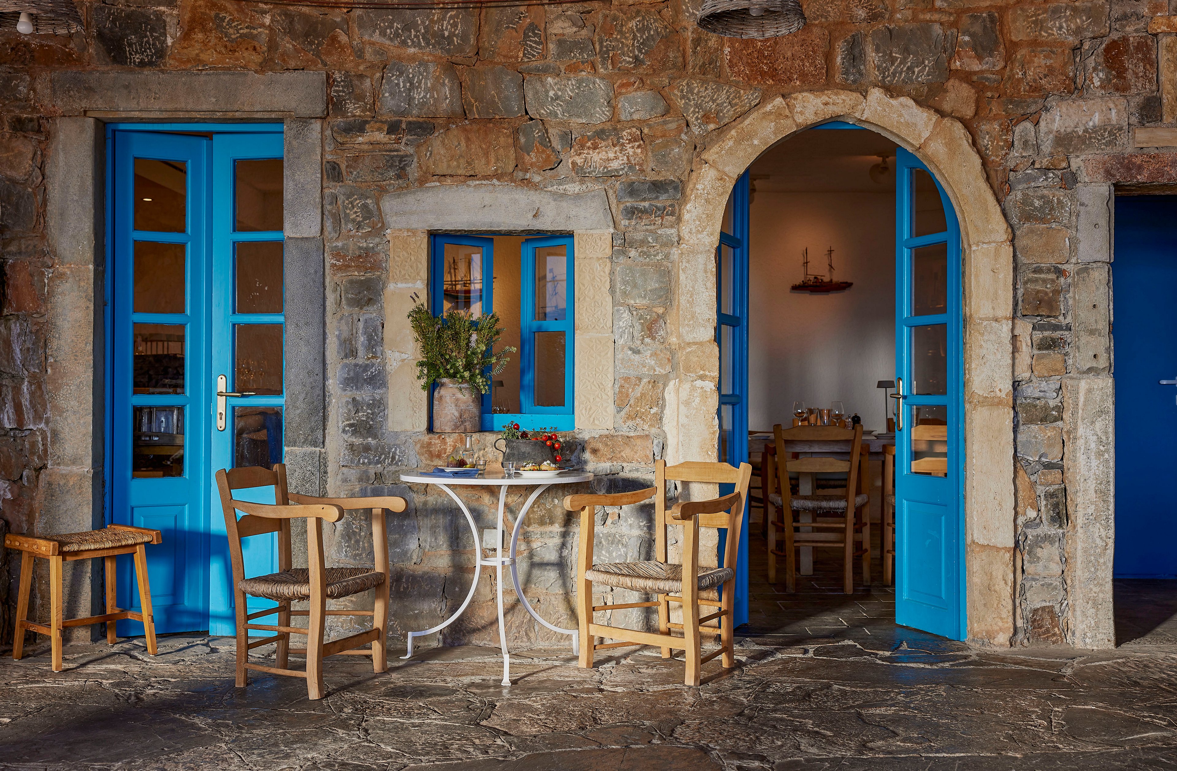 A small outdoor café is set against a rustic stone wall with vivid blue doors and window frames. A round white metal table with wooden chairs sits on a textured stone floor, creating an inviting seating area. Warm interior lighting and simple wooden furniture are visible through the open doorway, suggesting a relaxed dining space. The scene evokes a Mediterranean village atmosphere with charming, colorful details.