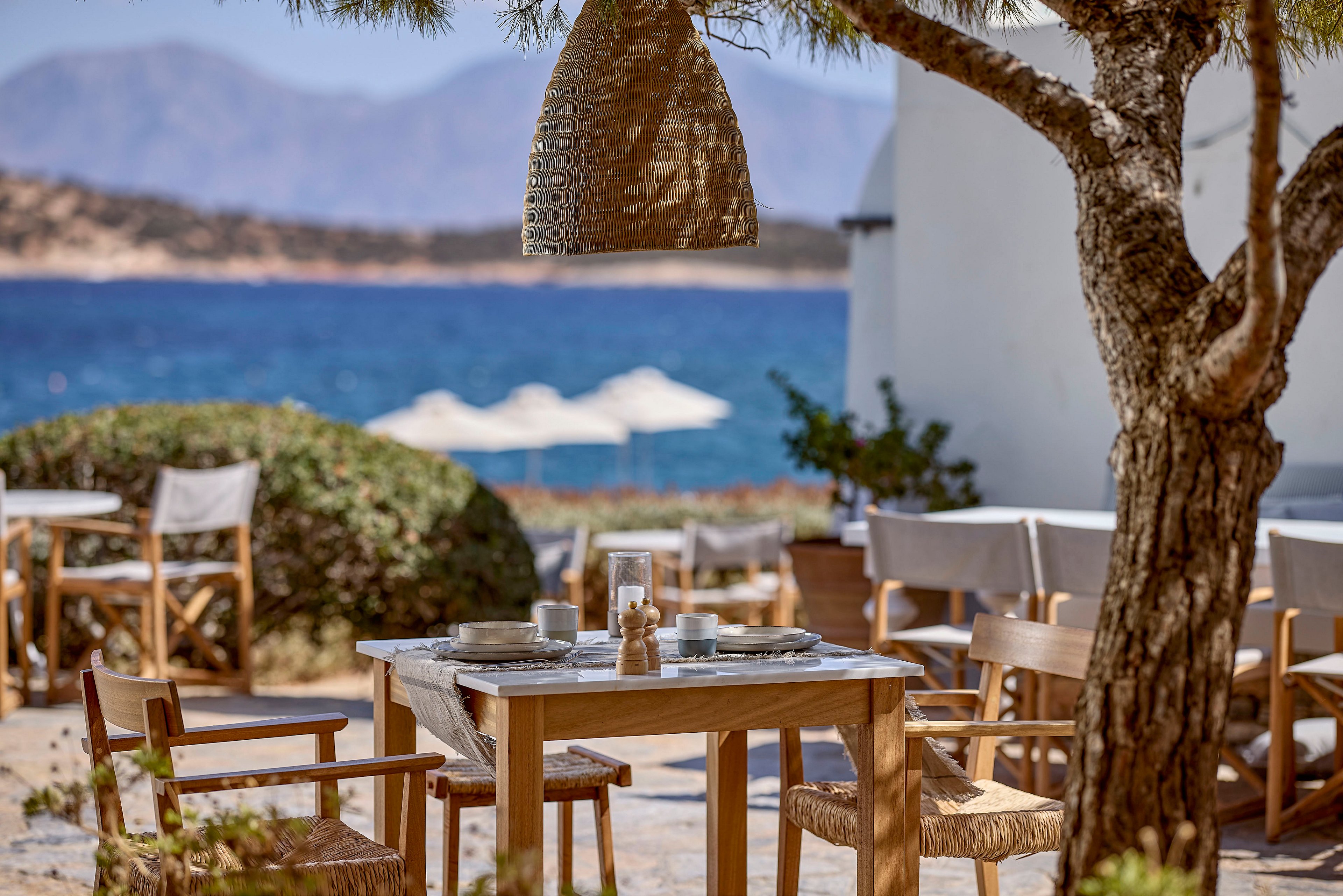 A wooden dining table is set with plates, glasses, and condiments in an outdoor café overlooking a calm blue sea. Wooden chairs surround the table, shaded partly by a tree and a woven pendant lamp. In the background, white umbrellas and additional seating areas face the water, with hazy mountains rising in the distance. The warm natural tones of wood and wicker contrast with the cool blues of the ocean and sky, creating a relaxed Mediterranean atmosphere.