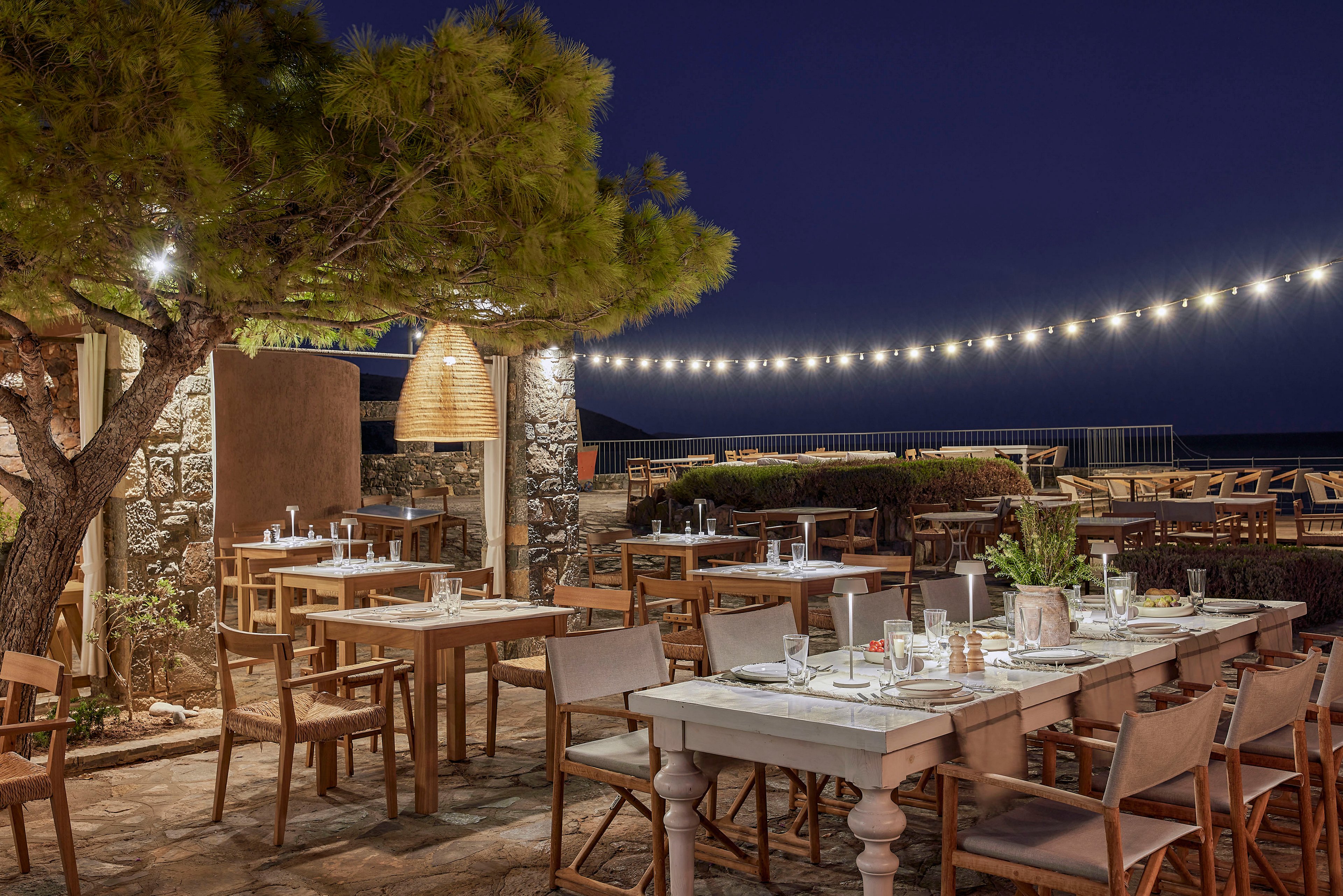 An inviting outdoor restaurant terrace is set for dinner under a deep blue night sky. Wooden tables and chairs are neatly arranged with glassware, plates, and candles, creating a warm and welcoming atmosphere. String lights hang overhead, adding soft illumination to the scene, while a large woven pendant lamp and nearby trees enhance the rustic charm. In the background, the sea and distant seating area suggest a scenic coastal location.