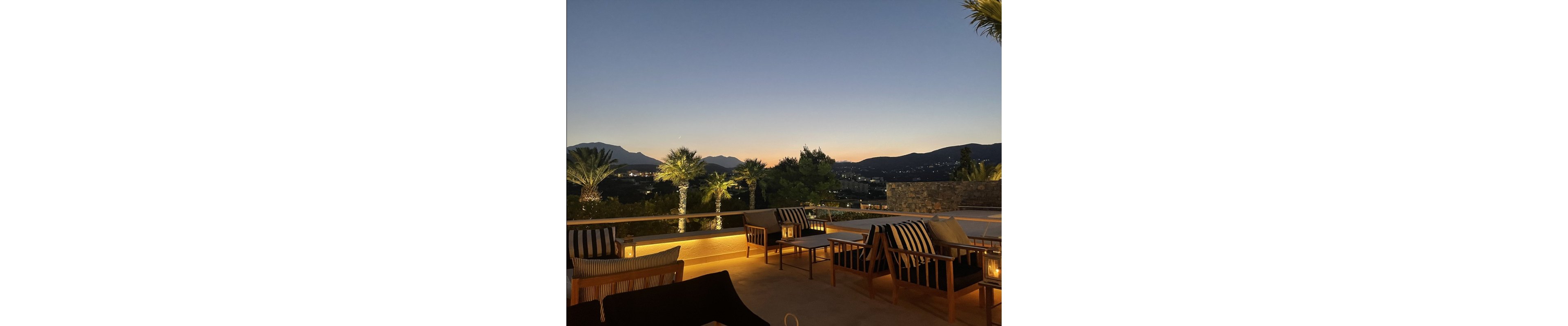 An outdoor terrace lounge is captured at dusk with comfortable wooden chairs and striped cushions arranged around small tables. Soft candlelight glows from lanterns, creating a warm and inviting atmosphere. Palm trees line the edge of the terrace, silhouetted against a clear evening sky. In the distance, hills and scattered lights from buildings are visible, adding depth to the tranquil scene.
