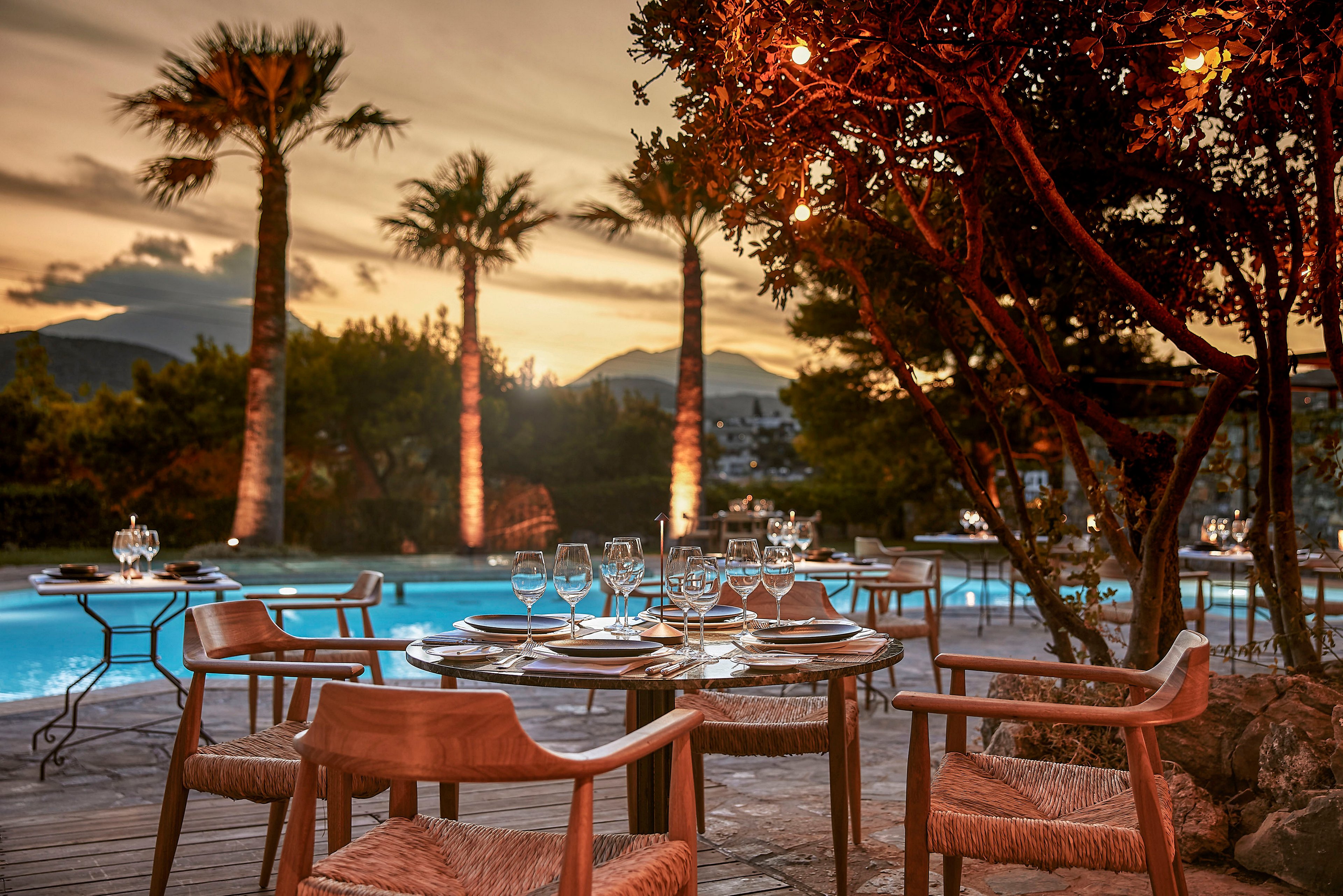 An outdoor restaurant table is set for dinner beside a glowing swimming pool at sunset. Wooden chairs with woven seats surround the table, which is arranged with plates, cutlery, and multiple wine glasses. Palm trees and lush greenery frame the scene, softly lit by warm accent lighting. Mountains are visible in the background, adding a tranquil, luxurious atmosphere to the evening setting.