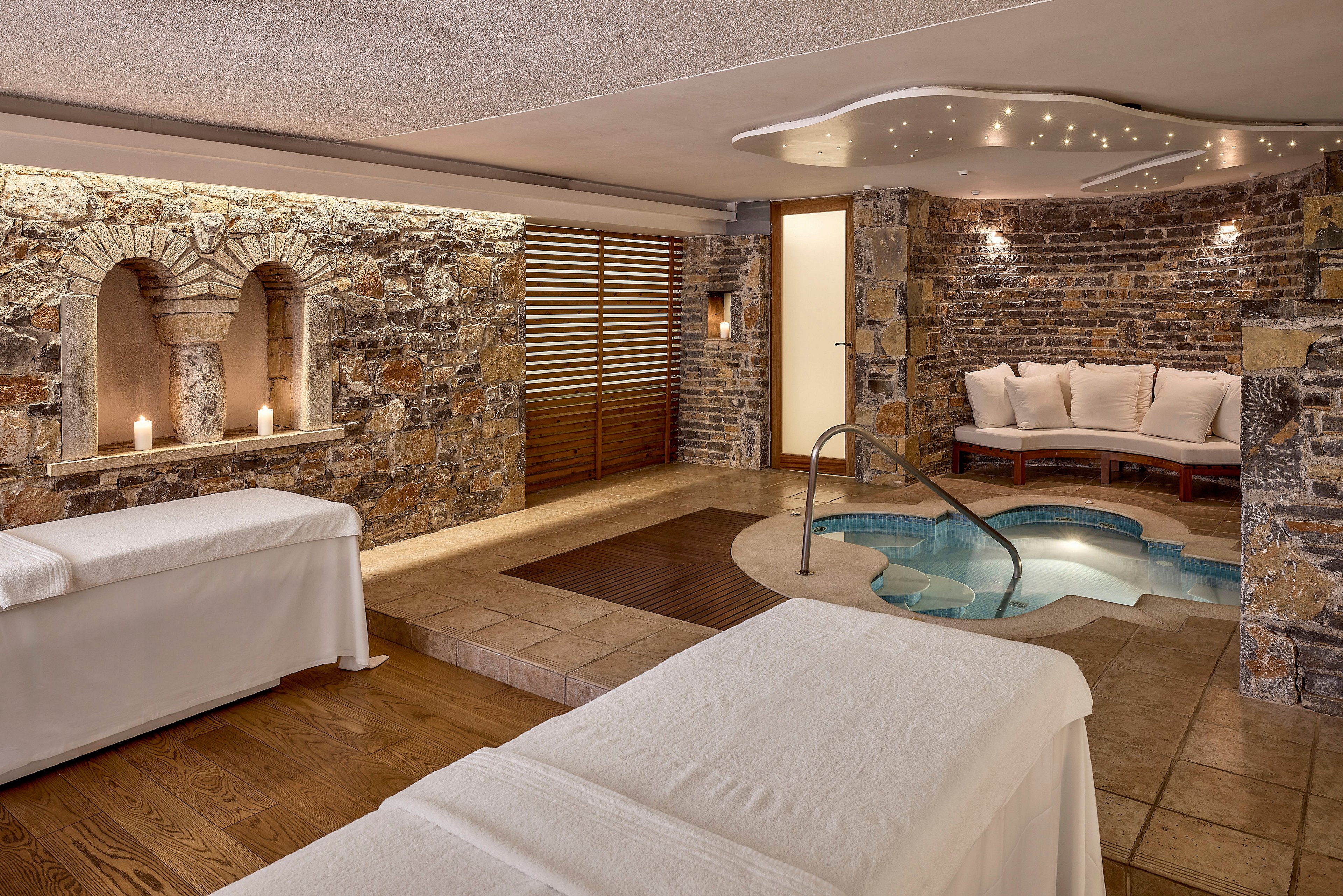 A serene spa room features two massage tables in the foreground, covered in crisp white linens. In the background, a small indoor pool with metal handrails is set into a tiled floor, surrounded by rustic stone walls. Soft lighting, recessed candles, and a cushioned seating area create a warm and inviting atmosphere. The overall scene suggests relaxation, wellness, and a high-end spa experience.