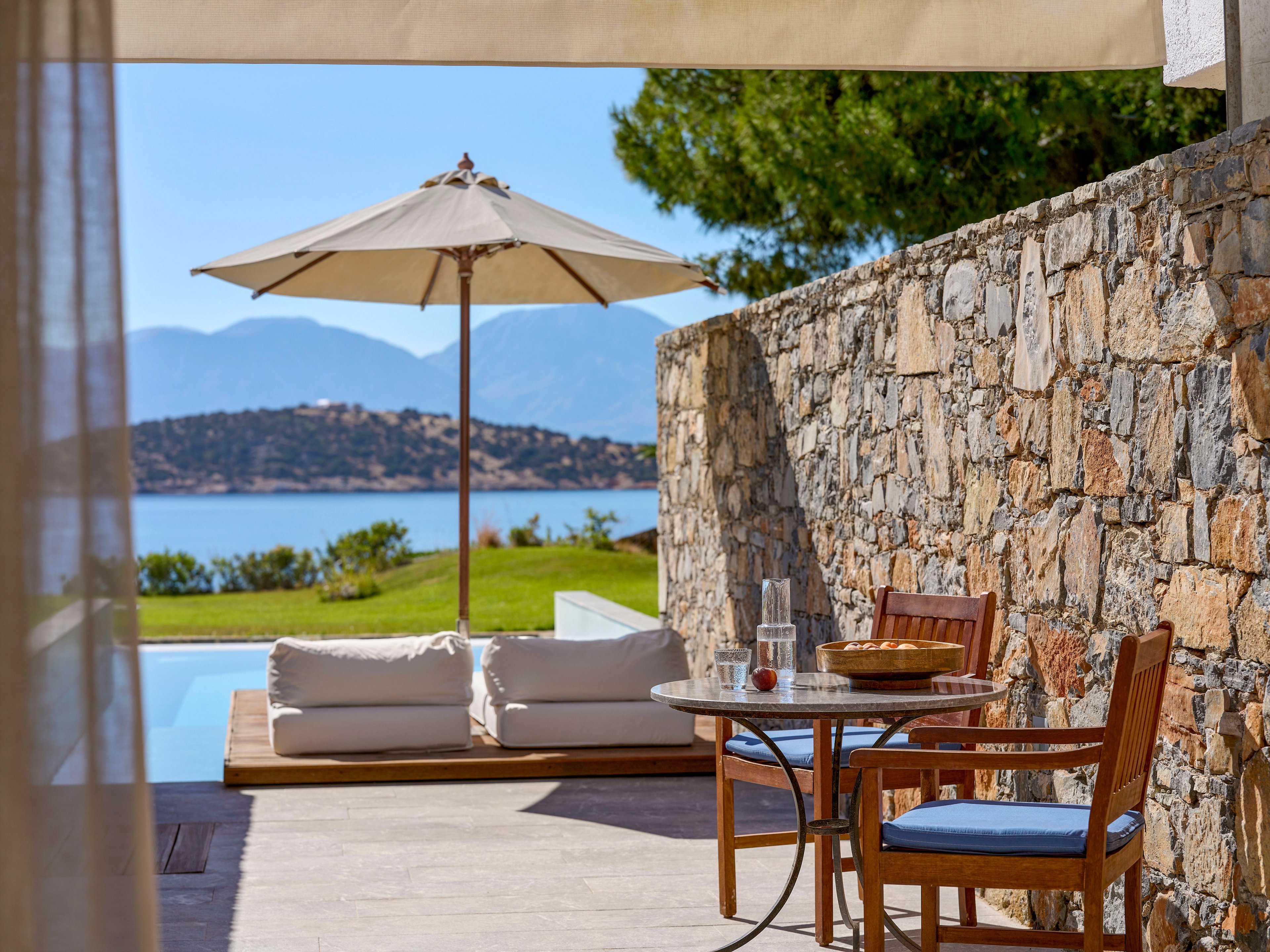 An elegant outdoor terrace features a small round table with wooden chairs beside a stone wall. In the background, cushioned sunbeds and a large patio umbrella sit next to a serene swimming pool. Beyond the pool, a grassy lawn leads to a calm blue sea with distant mountains under a clear sky. The scene suggests a peaceful vacation setting with bright natural light and warm, earthy tones.