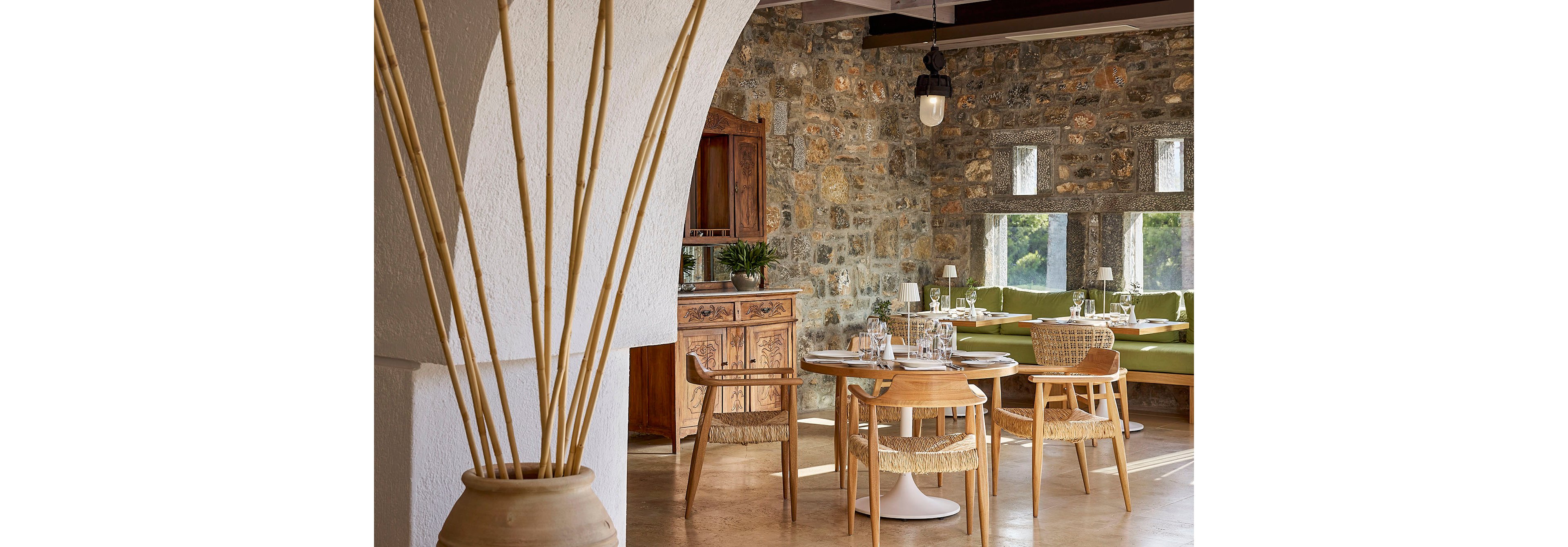 Inviting restaurant interior features wooden tables and woven chairs set against rustic stone walls. Natural light enters through small rectangular windows, illuminating green cushioned bench seating along the wall. A wooden sideboard and decorative vase with tall reeds add warmth and texture to the space. The overall atmosphere is relaxed and cozy, blending natural materials with contemporary dining decor.