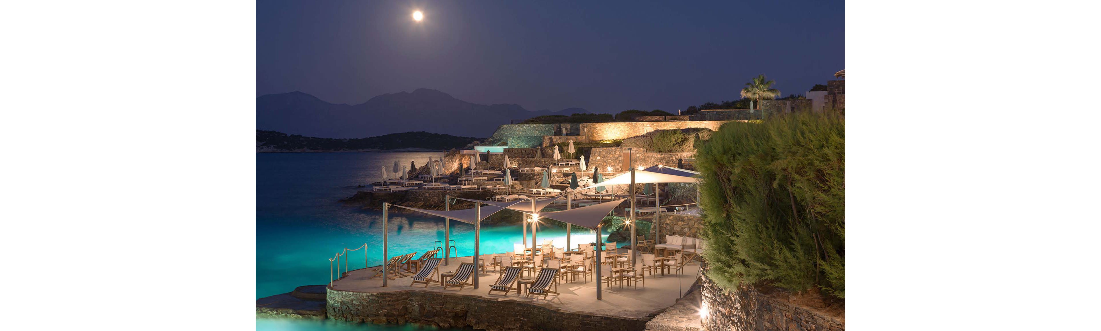 A coastal resort terrace is illuminated at night under a bright full moon. Lounge chairs and dining tables are arranged on a stone platform beside calm turquoise water. Soft artificial lighting highlights the seating area, umbrellas, and surrounding stone walls. Mountains and distant shoreline are visible in the background, adding depth to the tranquil seaside scene.