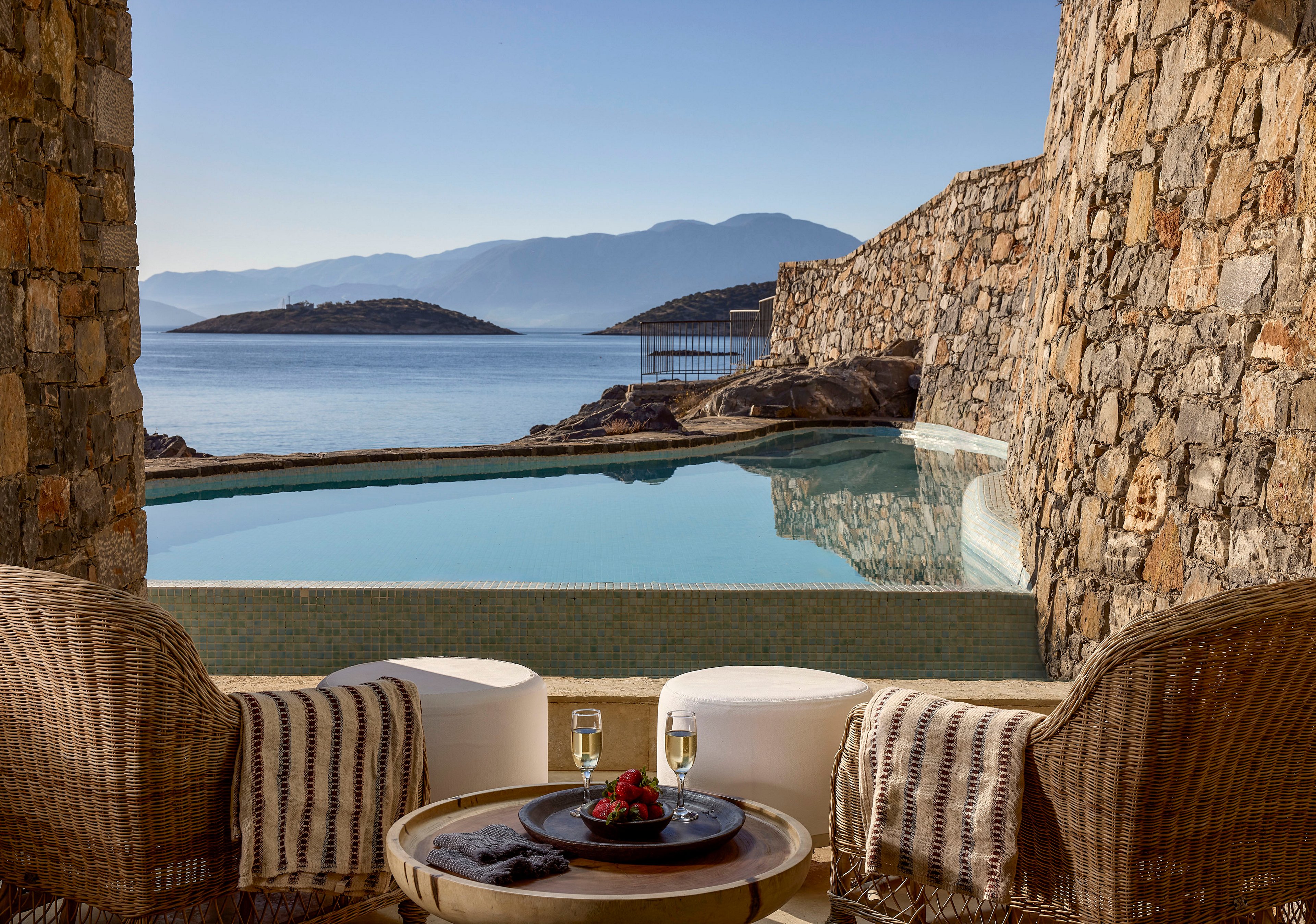 A private terrace overlooks a calm infinity pool facing a serene coastal landscape and distant islands. Two wicker armchairs and round stools surround a small table set with dessert and glasses of white wine. Stone walls frame the view of the blue water and clear sky, creating an intimate, luxurious atmosphere. The scene suggests a peaceful vacation retreat by the sea.